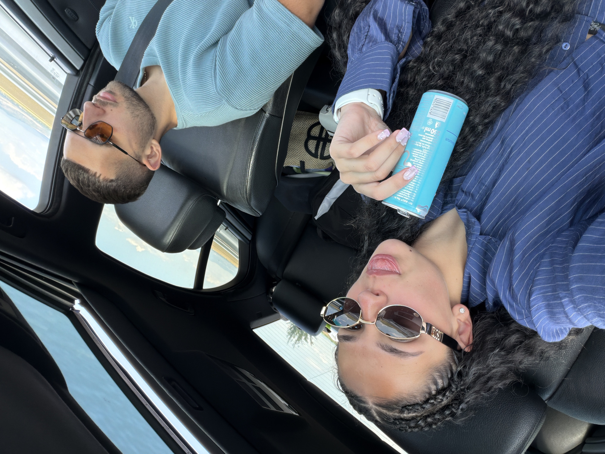 The image shows two individuals inside a car. The person in the foreground is a woman with long, dark, curly hair, wearing a blue pinstripe shirt and sunglasses. She is holding a blue can in her right hand. She appears to be sticking her tongue out slightly. The person in the background is a man wearing a light blue, ribbed t-shirt and sunglasses. He is looking upwards with his eyes closed. The car has a sunroof that is open, allowing natural light to enter. The interior of the car appears to be black leather. The setting appears to be during daylight, with a glimpse of the sky and a landscape that suggests a rural or coastal environment outside the car. There is no indication of the location being Polsko Padarevo, Bulgaria.
