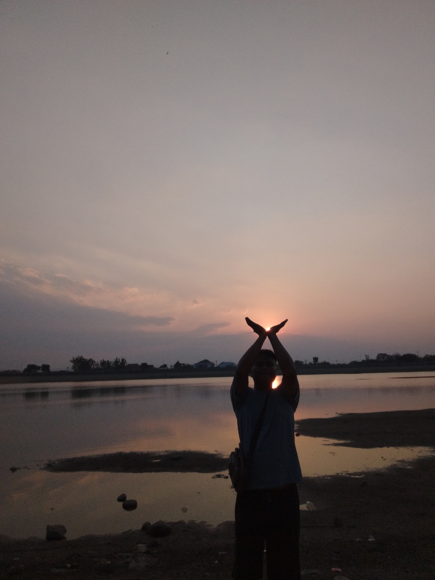The image depicts a silhouette of a person standing on the shore of a body of water, likely a lake or river, at sunset. The person is holding their hands up and together, framing the sun which is partially obscured by their palms. The sun appears as a bright orb, casting warm hues of orange and yellow across the sky and reflecting on the water's surface. The sky is a pale pink and grey, with wispy clouds. In the background, a line of trees and some structures are visible on the distant horizon, suggesting a rural or semi-urban setting. The foreground shows a dark, sandy shoreline with a few rocks. The scene appears to be peaceful and serene, capturing a moment of natural beauty. Given the context provided, this scene could be located in Sambirejo, Indonesia, a region known for its natural landscapes.