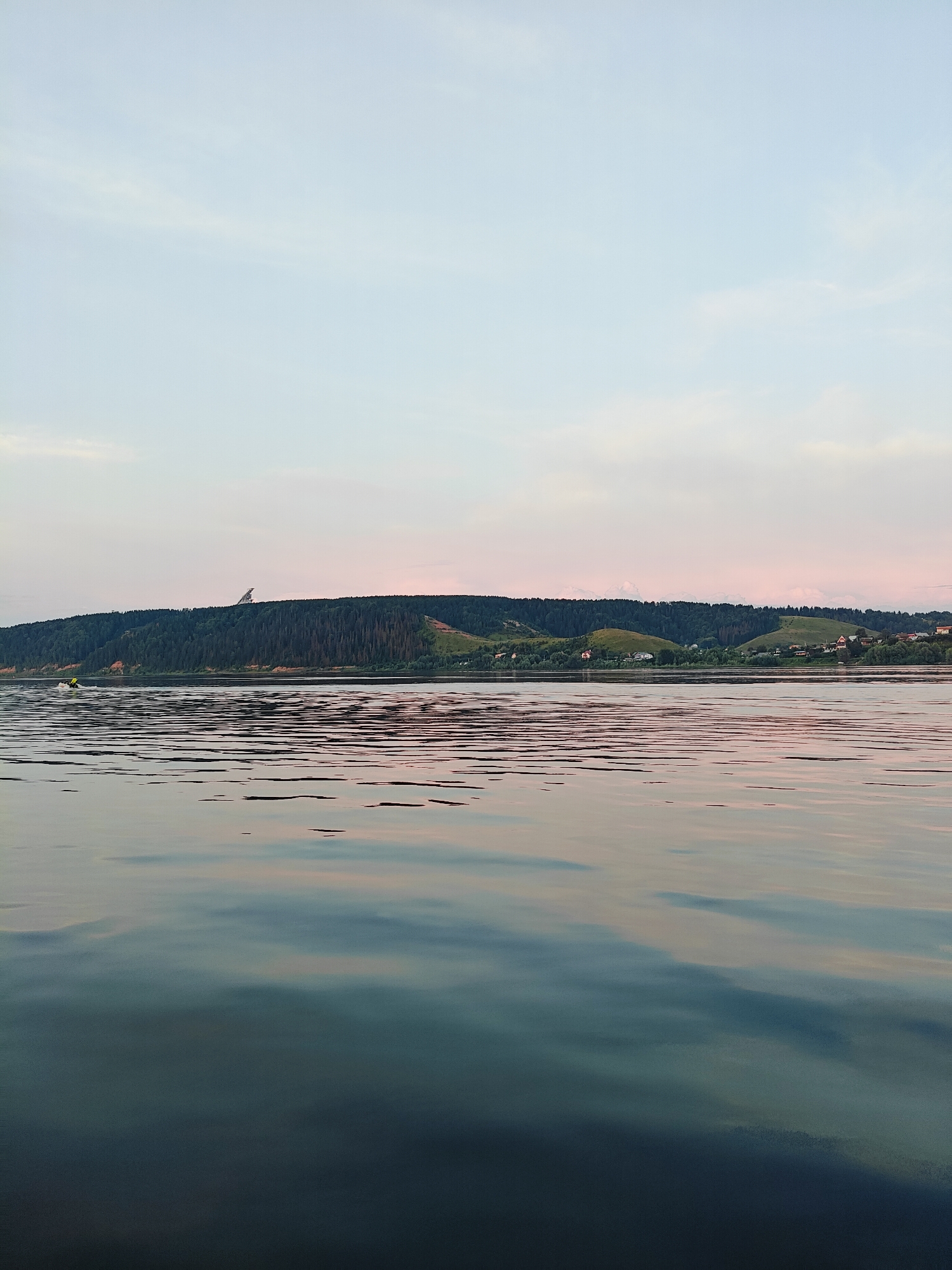 The image depicts a calm body of water, likely a river or lake, with a forested hill in the background. The sky is a pale blue with subtle pink clouds. A small, yellow object, possibly a boat or a person on a personal watercraft, is visible on the left side of the water. The water's surface reflects the colors of the sky and the landscape. The hill in the background is covered in dense green trees and features some scattered houses and a distinctive, tall, angular structure on its crest. The overall scene suggests a peaceful, natural setting at dusk or dawn. The location is identified as Noviy, Russia.