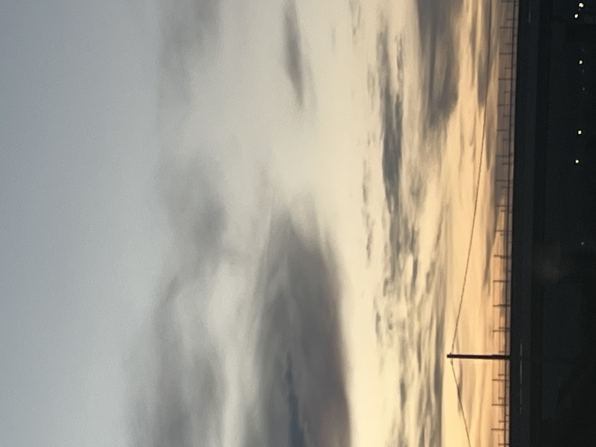 The image depicts a cloudy sky at sunset or sunrise in Hrabarsko, Bulgaria. In the foreground, there's a silhouette of a structure, possibly a building or bridge, with a pole or mast extending upwards, bisecting the sky. A power line is also visible. There are some lights in the distance.