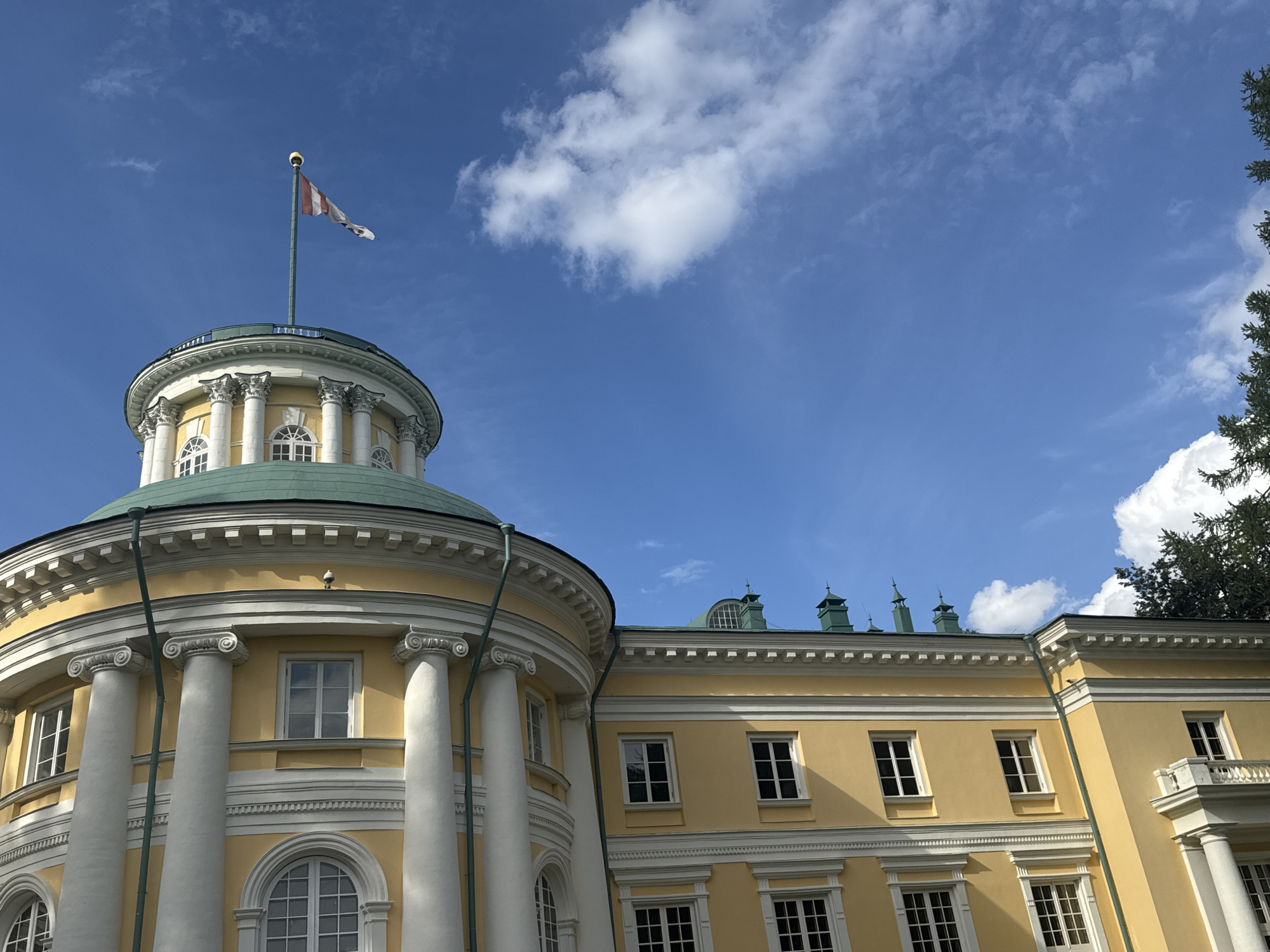 The image shows a bright yellow building with white architectural details under a clear blue sky with scattered white clouds. A flagpole with a white and red striped flag is visible on top of a domed section of the building, which is supported by large white columns. The building has several windows, some arched and some rectangular. To the right of the domed section, a longer section of the building with a green roof and multiple smaller green dormer windows is visible. The overall architectural style appears classical or neoclassical. The scene is likely outdoors, with a glimpse of dark green foliage from a tree on the far right edge of the image. The location is the Arkhangelskoye Estate Museum-Reserve, located in the Moscow Oblast, Russia, near Moscow.