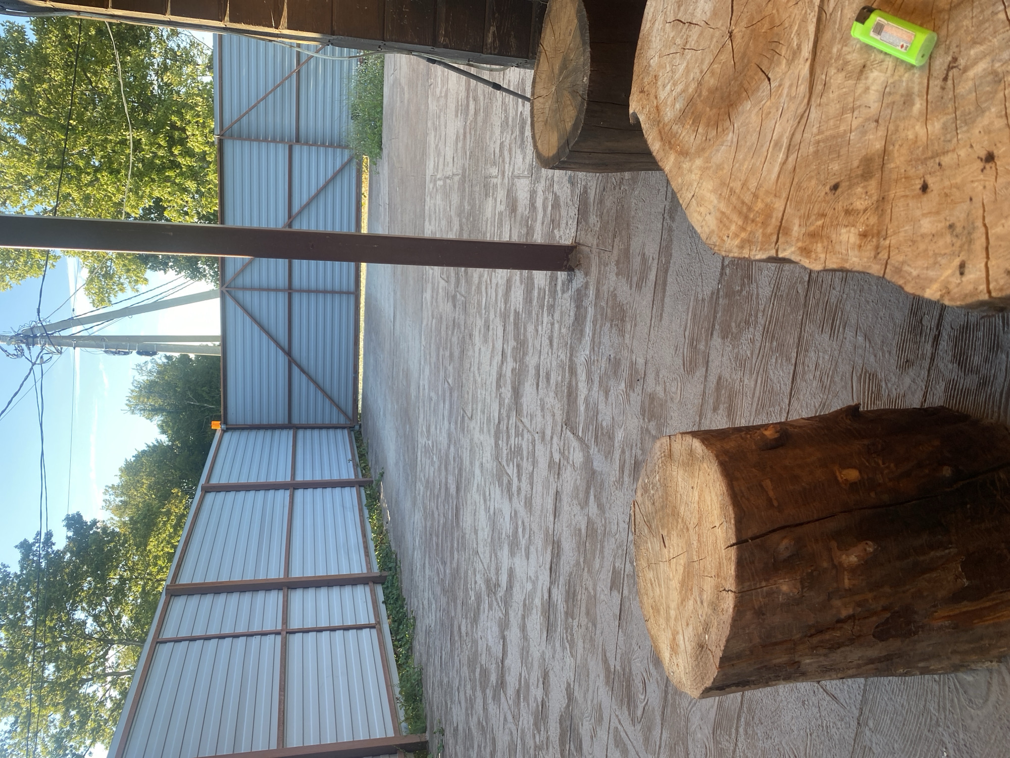 The image depicts a backyard or outdoor patio area with a light-colored, patterned concrete floor. A metal fence, likely corrugated, borders the area, with wooden posts supporting it. There are two large, rough-hewn tree stumps in the foreground, one to the left and another to the right, serving as seating or decorative elements. A bright green lighter rests on the surface of the tree stump on the right. In the background, trees and utility poles are visible under a clear blue sky. The overall impression is of a rustic, outdoor space. The location is stated to be the city of Aderbievka, Russia.
