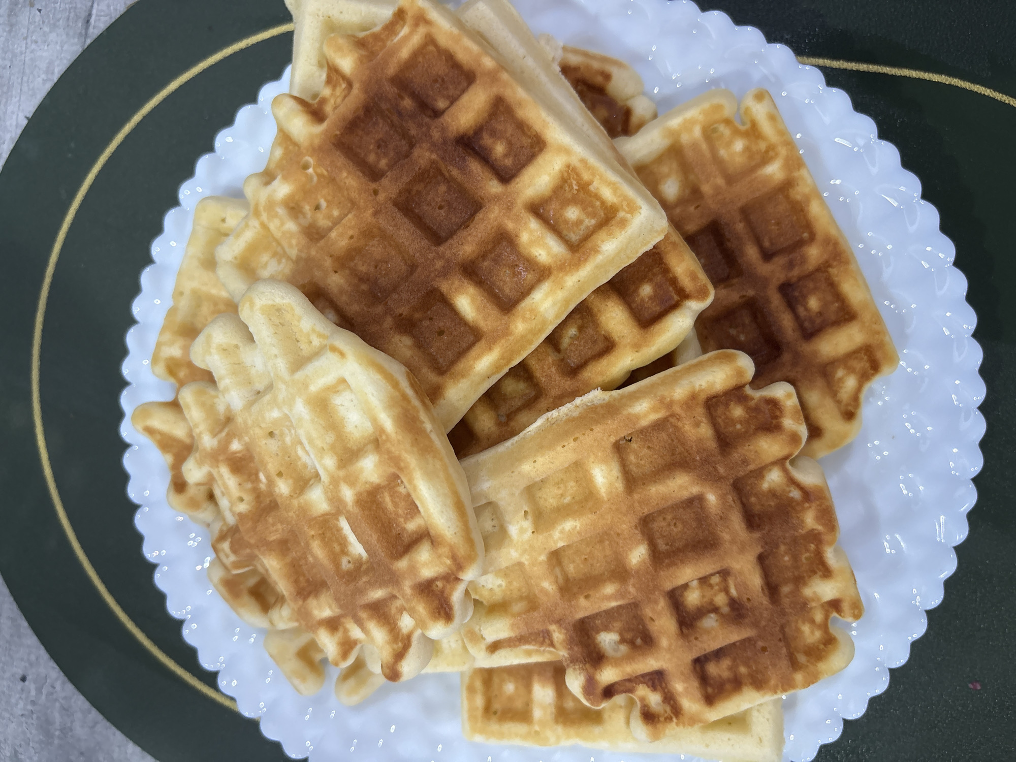 The image presents an overhead, close-up view of a stack of approximately ten to twelve golden-brown waffles, situated on a white, scalloped-edge plate in Chaikovsky, Russia. Each waffle exhibits a distinct grid pattern, with color variations ranging from lighter golden to darker toasted brown. The waffles are irregularly stacked. The white plate rests upon a dark green surface, which displays a thin, curved golden line. A light grey, textured surface is visible in the upper left background. No individuals, actions, or interactions are depicted within the frame.