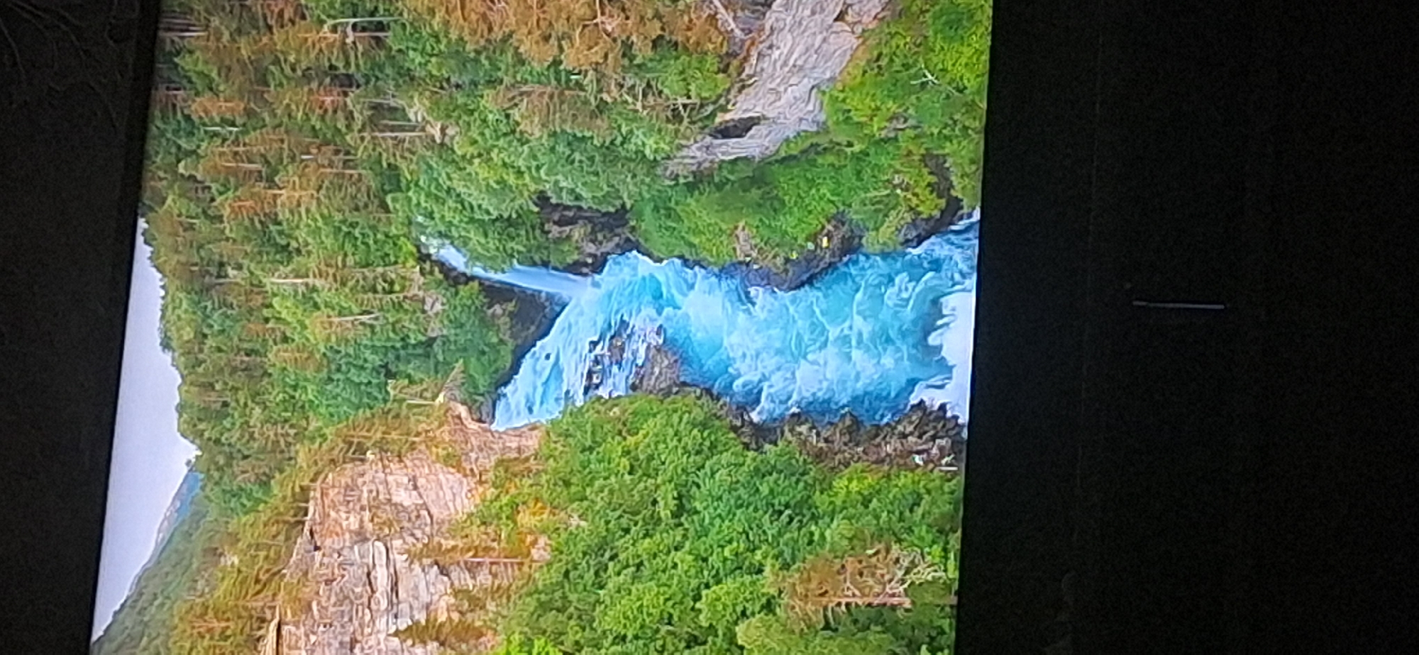 The image depicts a scenic view of a powerful river cascading through a lush, green canyon. The water is a vibrant blue, indicating its clarity and speed, with white frothy rapids visible. Towering cliffs flank the river, covered in dense evergreen trees. The sky above is a pale gray. The scene is an example of natural beauty found in mountain or forest regions. The provided crops offer closer views of the forested slopes, rocky cliffs, and the turbulent river. The image is presented on a dark surface, likely a screen, with a slight glare. There is no indication of the city of Bila Tserkva, Ukraine, in the image.