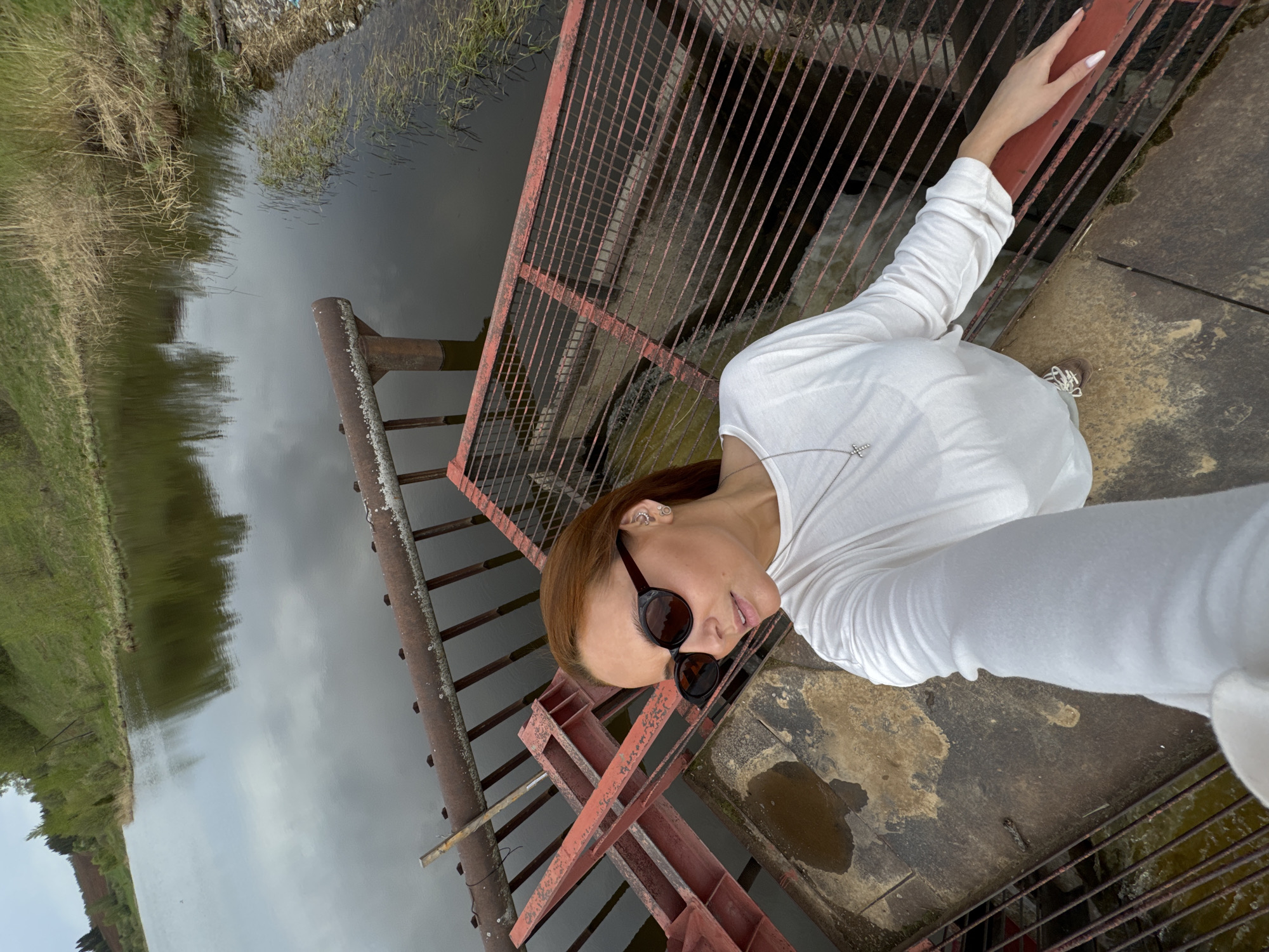 A woman wearing sunglasses and a white long-sleeved shirt stands on a rusty metal structure with a red grid, overlooking a body of water. The water reflects the cloudy sky. Green vegetation is visible on the banks. The location is likely Nozhovka, Russia.