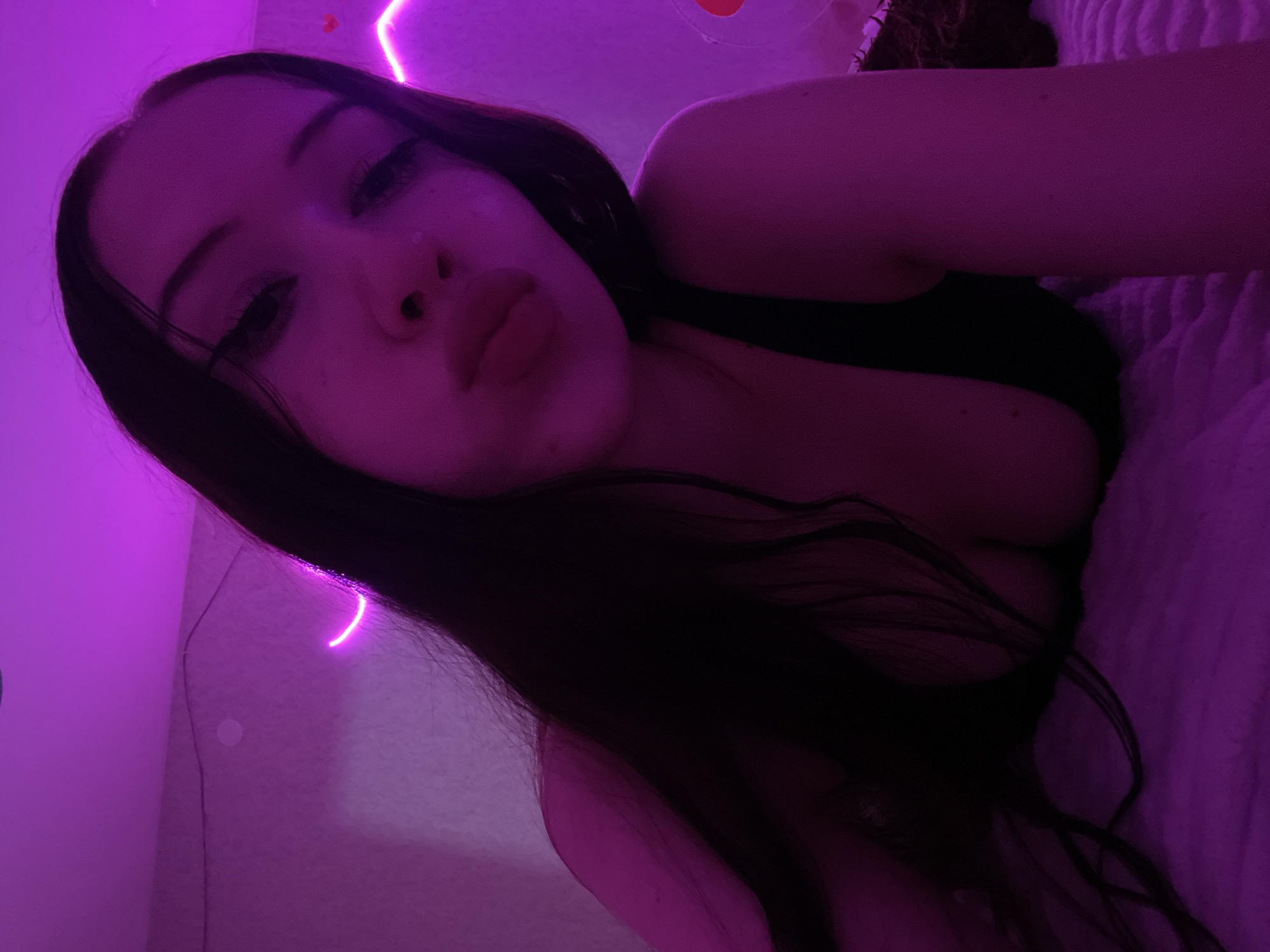 A young woman is lying on a bed, illuminated by purple neon lights. She is looking towards the camera with her lips pursed. Her dark hair is spread around her face and shoulders. The background is a wall with a pink neon light strip. The overall atmosphere is intimate and dimly lit. The exact location in Votkinsk, Russia, is not identifiable from the image.