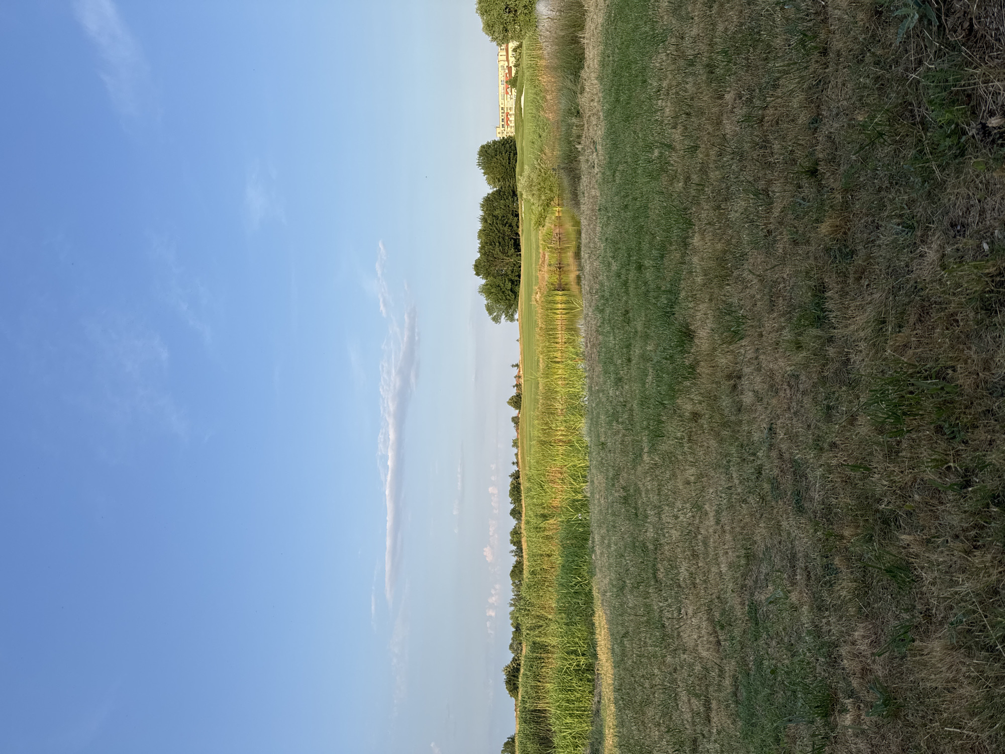 This is a landscape photograph of a golf course in Balchik, Bulgaria. The foreground is a dry, grassy field. Beyond this is a body of water with reeds growing along its edge. In the middle ground, there is a lush green golf course with trees and a building in the background. The sky is blue with a few wispy clouds.