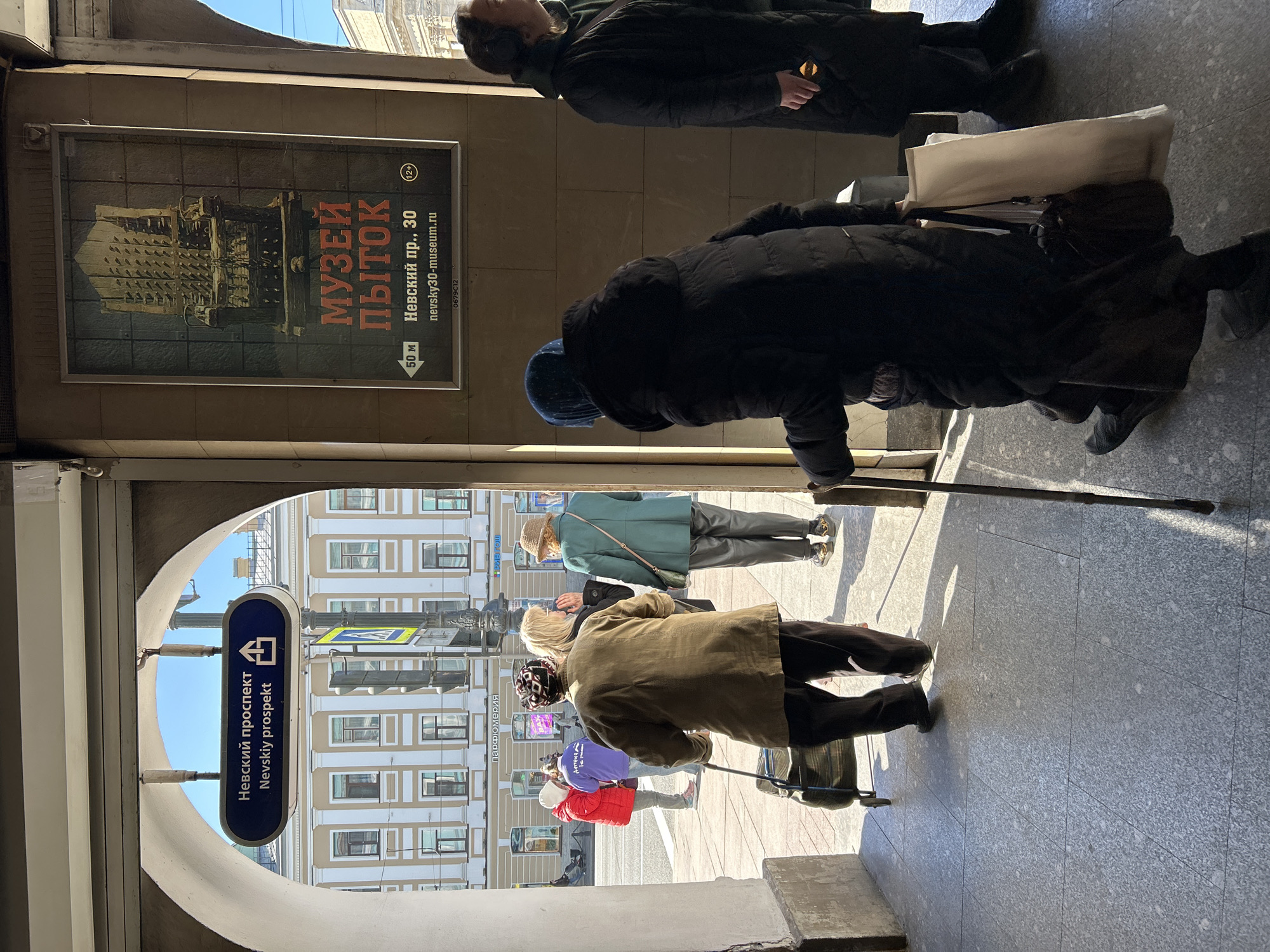 This photograph shows a partially obscured view of a street scene through an archway, likely the entrance to a subway or underground passage. On the left side of the archway, there is a sign indicating the location as "Nevskiy prospekt" (Nevsky Prospekt) in both Russian and English, thus indicating the location is Saint Petersburg, Russia.
Several people are visible within the frame, with some standing inside the passage and others walking on the street outside. A person wearing black clothes and headphones stands in the passageway. An elderly woman with a walking stick is also visible. Additional pedestrians are walking on the street outside.
A poster advertises the "Museum of Torture" and provides its address as "Nevskiy пр., 30" (Nevsky Prospekt, 30) and website, nevsky30-museum.ru. The poster also has a "12+" age restriction warning. The sign indicates the museum is 50 meters away.