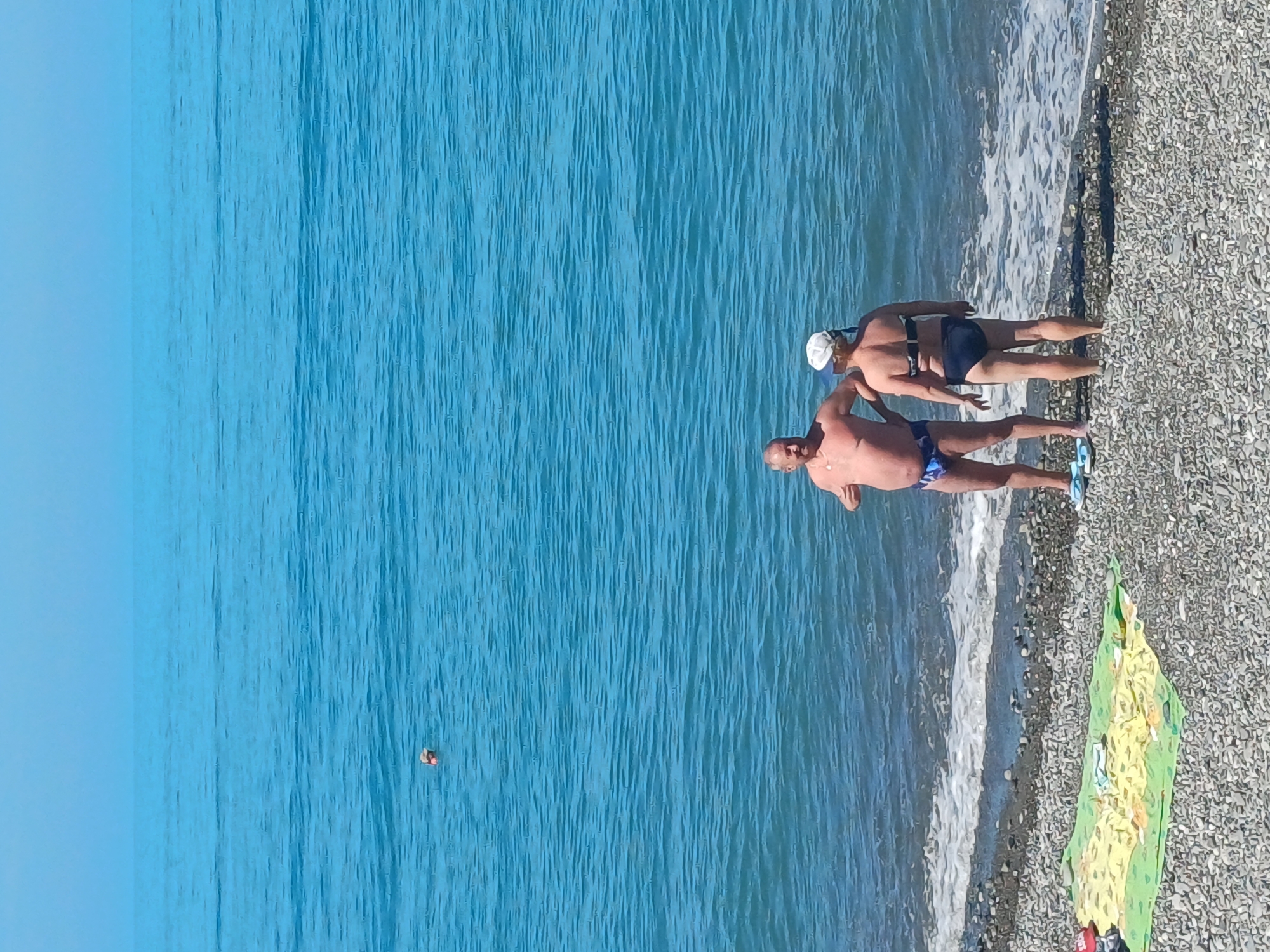 Two individuals, a man and a woman, stand on a pebble beach by the Black Sea in Sochi, Russia. The man, with a shaved head and wearing blue swim trunks, is facing the woman. The woman, wearing a white cap and a dark bikini, has her back to the viewer. The sea is calm with gentle waves lapping the shore. In the background, a small orange buoy floats in the water. A colorful towel is spread on the beach to the left of the couple. The sky is clear and blue.