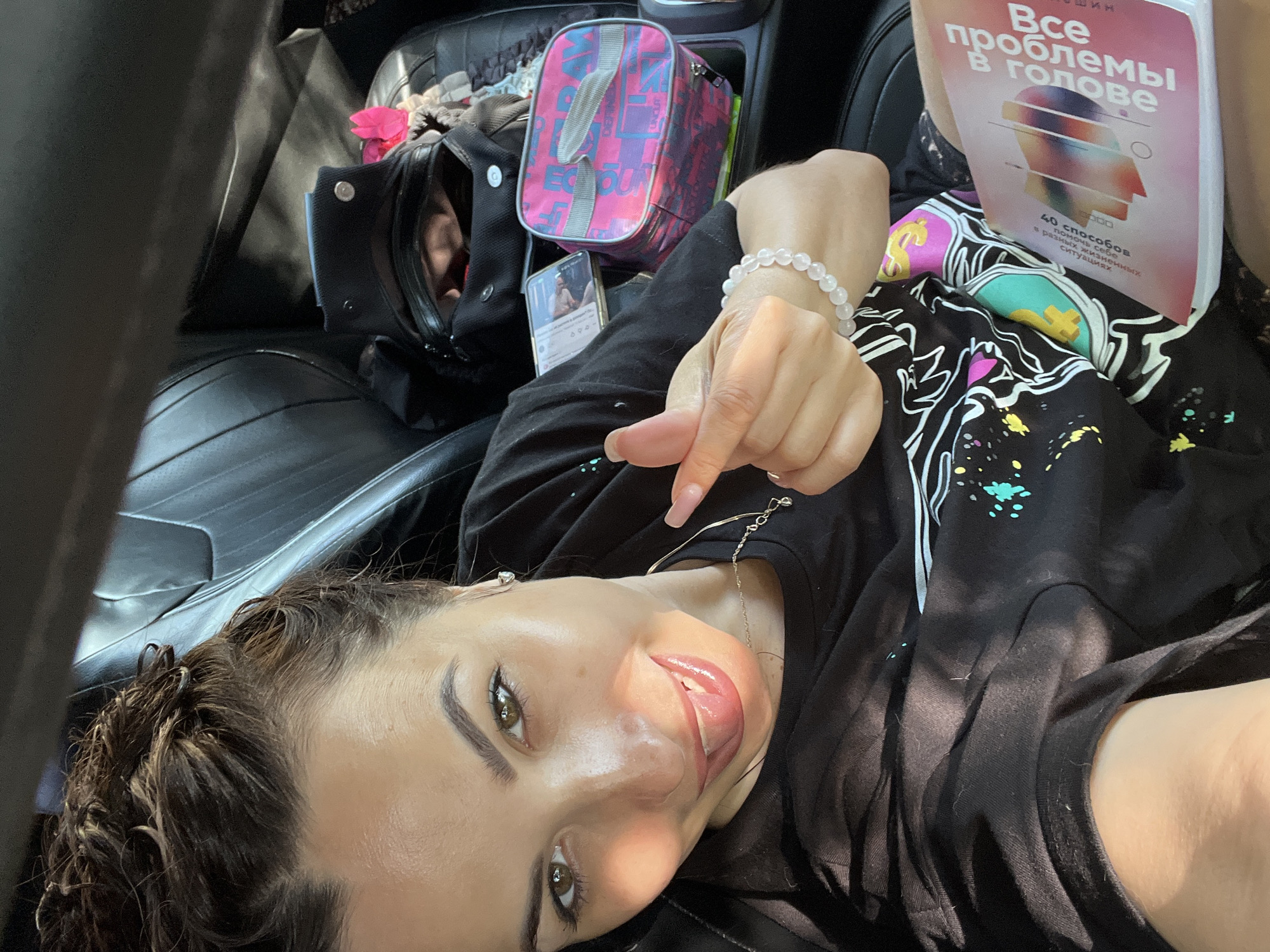A woman is pictured from the chest up, sitting in the driver's seat of a car. She is wearing a black t-shirt with a colorful graphic design. Her hair is pulled up in a bun. She is sticking her tongue out and making a hand gesture. She is holding a book titled "Все проблемы в голове" (All problems are in the head) in her right hand. Behind her, on the passenger seat, is a black bag and a pink lunchbox. The car's interior appears to be black leather.
