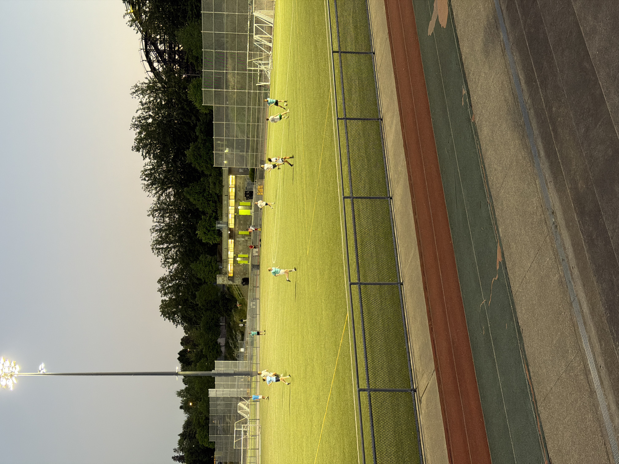 An artificial turf soccer field is illuminated by tall light poles during dusk. Several individuals are playing soccer on the field. In the background, a dense line of trees separates the field from a roller coaster track, and a building with illuminated windows is visible to the right. A chain-link fence encloses the field, and a running track with red and green lanes is in the foreground. The scene is consistent with a public park or school sports facility in Vancouver, Canada.