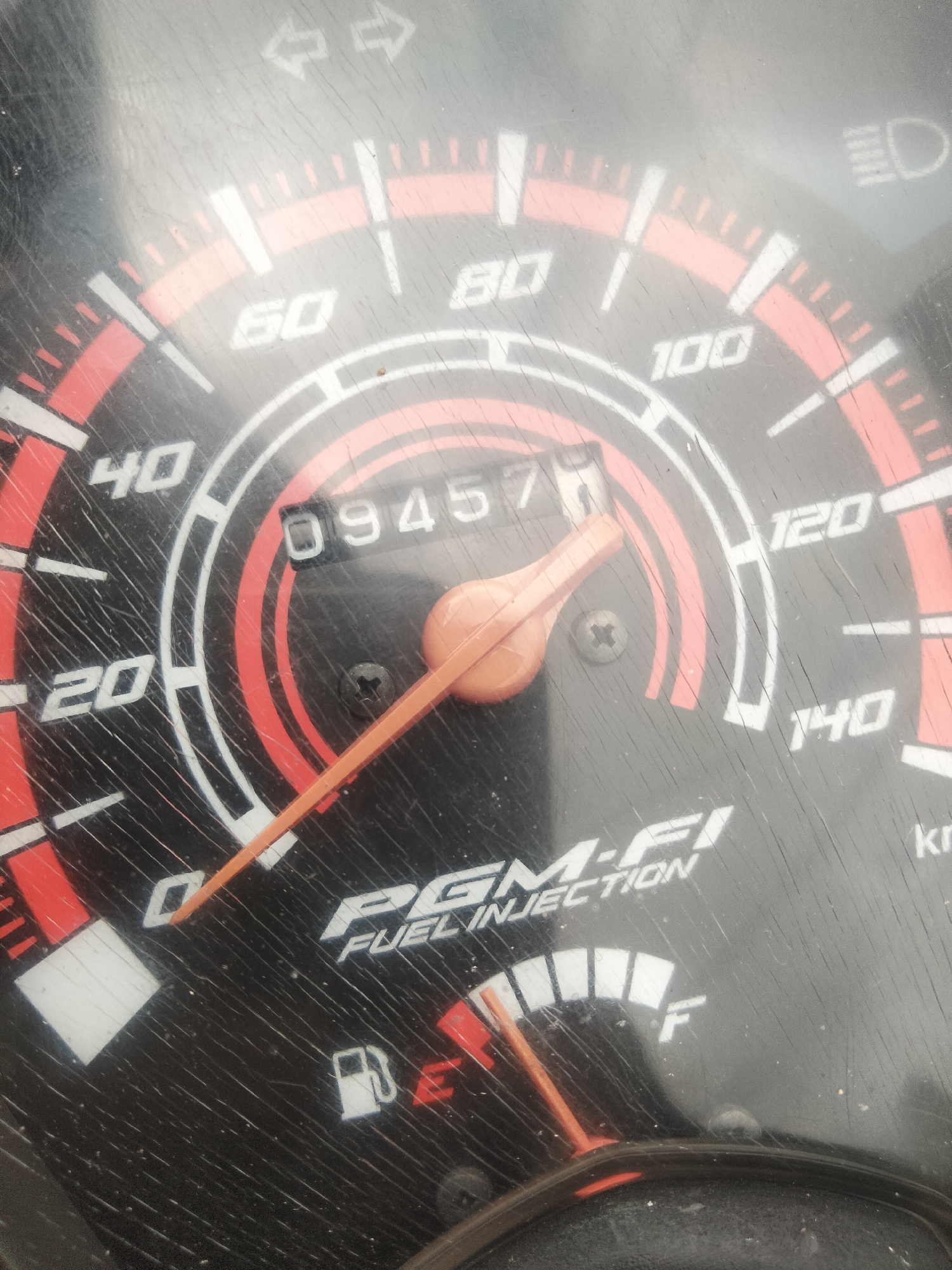 The image is a close-up of a motorcycle's speedometer. The speedometer has markings for speed in kilometers per hour, with numbers like 20, 40, 60, 80, 100, 120, and 140 visible. There is also an odometer displaying the number "09457". A fuel gauge is partially visible, indicating the fuel level. The speedometer face is dusty and scratched. The PGM-FI FUEL INJECTION text is visible, indicating the motorcycle uses fuel injection. The image appears to be taken in Ketanggung, Indonesia, though no specific location details are visible in the photograph itself.