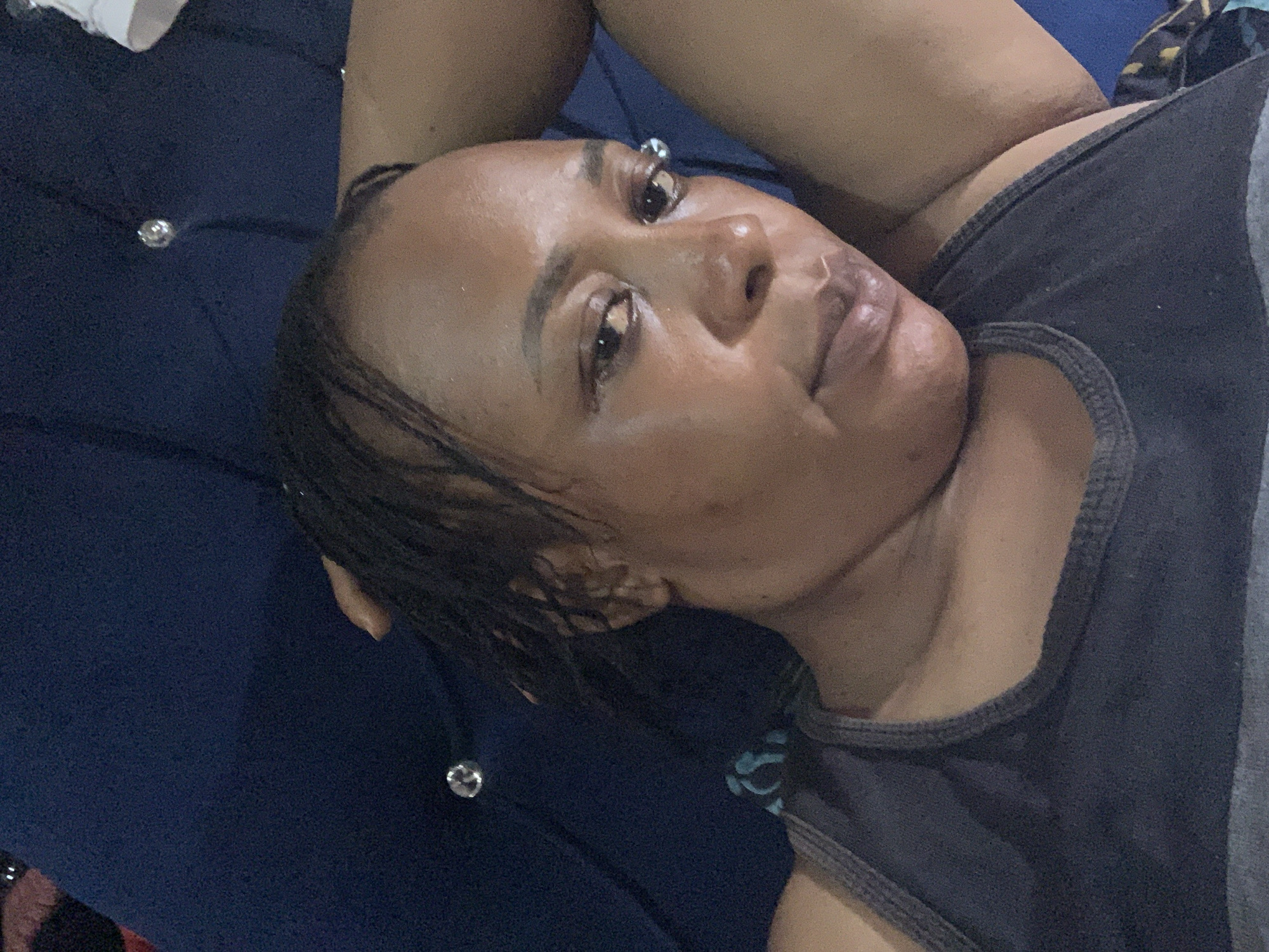 The media file shows a close-up, overhead view of a person lying down on a dark blue, tufted surface. The person, likely a woman, is lying on her back, with her head tilted to the right. She has dark hair, which appears wet and is slicked back. Her eyes are open and looking upwards. She is wearing a dark tank top with grey trim. The surface she is lying on has several clear, faceted buttons or studs, likely part of the upholstery.
The lighting suggests it might be indoors or in an area with artificial light, as there are no strong shadows indicating natural sunlight. The overall mood conveyed is one of relaxation or rest. There is no visible text in the image. The context provided places this scene in Sabon Tasha, Nigeria.