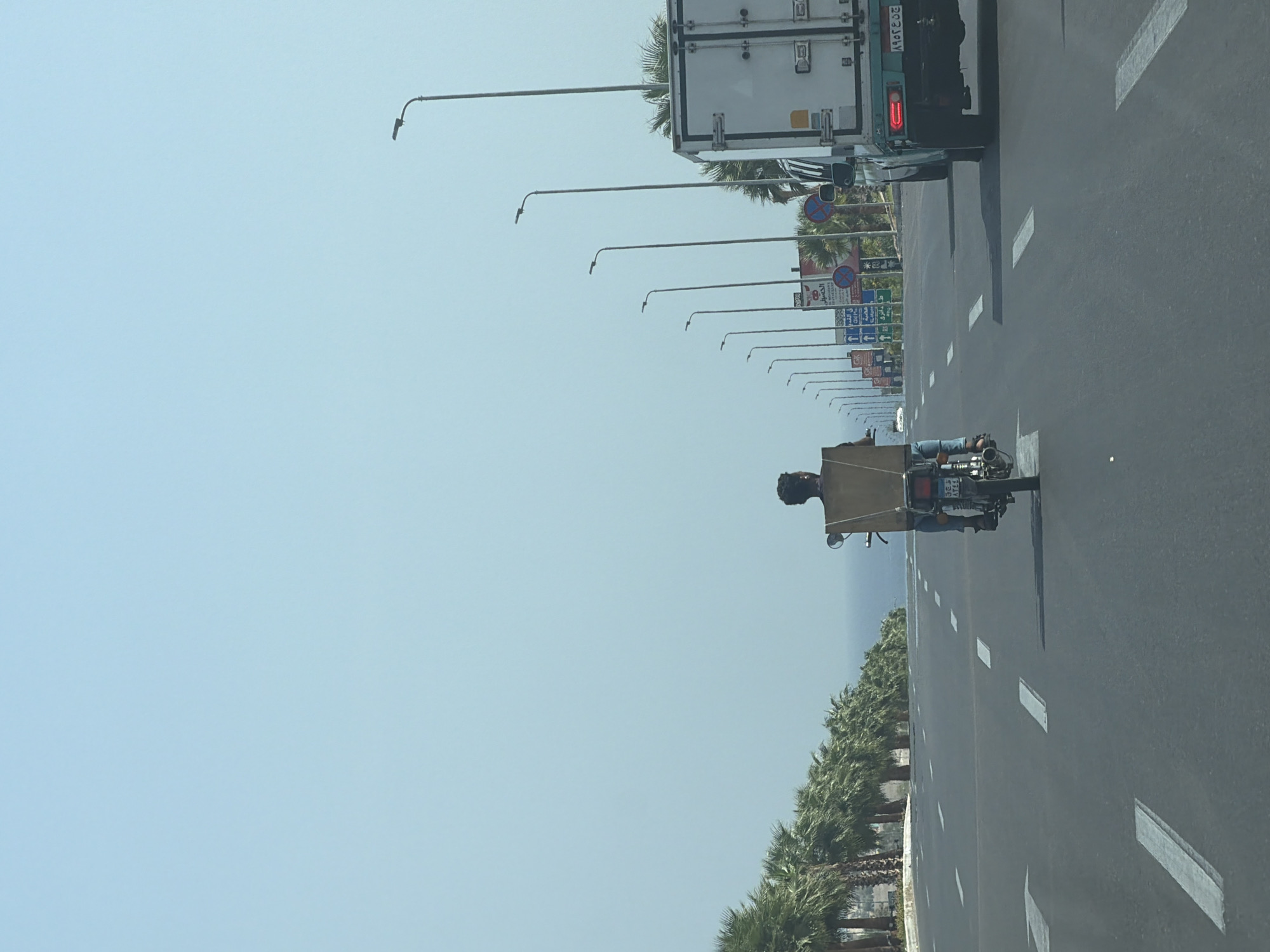 This image depicts a street scene in Sharm El Sheikh, Egypt, on a clear day with a pale blue sky. The foreground shows a paved road with white lane markings, indicating a multi-lane highway. In the center of the road, a person is riding a motorcycle, facing away from the camera. The motorcyclist is wearing blue jeans and a brown cardboard box is strapped to the back of the motorcycle, obscuring the rear of the rider. To the right of the motorcycle, a white delivery truck is visible, also moving forward. The truck has a license plate with Arabic characters and numbers.
The environment includes a line of palm trees on the left side of the road, suggesting a tropical or subtropical location. In the background, a body of water, likely the Red Sea, can be seen beyond the palm trees. Several streetlights are positioned along the right side of the road, and various road signs are visible in the distance, some with Arabic text, further confirming the location in Egypt. The overall impression is of a typical highway scene in a coastal city.