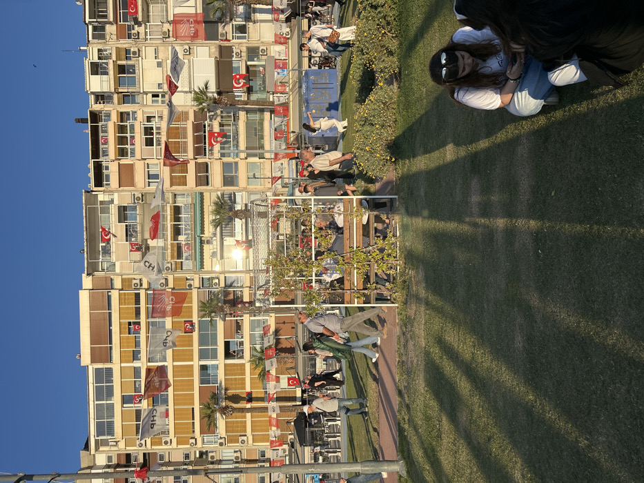 The image depicts a public park in İzmir, Türkiye, located at the coordinates 38.438 latitude and 27.141 longitude. The scene features a grassy area, a large, multi-story building, and a metal trellis structure. People are walking and interacting in the park. Some are seated on a bench within the trellis, while others are sitting on the grass. Shadows cast from the setting sun stretch across the grass. Several Turkish flags are draped across the building's facade.