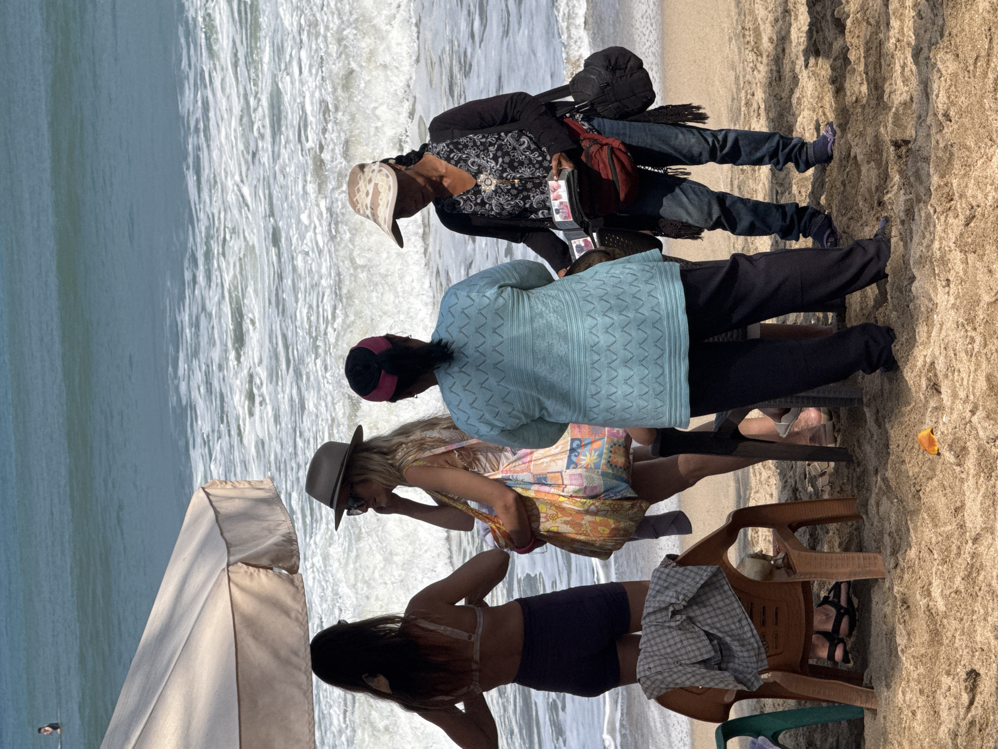 A group of four people are standing on a sandy beach in Kuta, Indonesia, with the ocean and waves in the background. One person, wearing a black top with a white floral pattern, a black open cardigan, and blue jeans, is wearing a camouflage visor and is holding a small black case with what appear to be photographs or small items for sale. She has a red fanny pack around her waist and a black bag slung over her shoulder. To her left, another person is wearing a light blue long-sleeved top with a textured pattern, and dark pants. To the left of these two, a woman with blonde hair, wearing a grey fedora and a colorful patterned dress, is holding a large patterned tote bag. She is looking towards the person in the fedora. The fourth person, on the far left, is wearing a bikini top and dark shorts and has her back to the camera, with her hair tied back. There is a beige beach umbrella providing shade to some of the group. In the foreground, there are two plastic chairs, one brown and one partially visible green chair, with clothing draped over the brown chair. The sand is a light brown color. The water is a turquoise blue with white waves breaking.