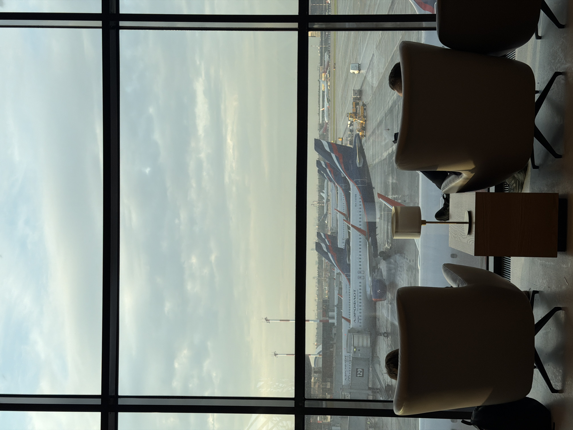 An indoor shot looking out a large window at an airport. Several airplanes sit on the tarmac. Two chairs are in the foreground, one is occupied by a person, with a table and a lamp between the chairs. The location appears to be a lounge area in an airport.