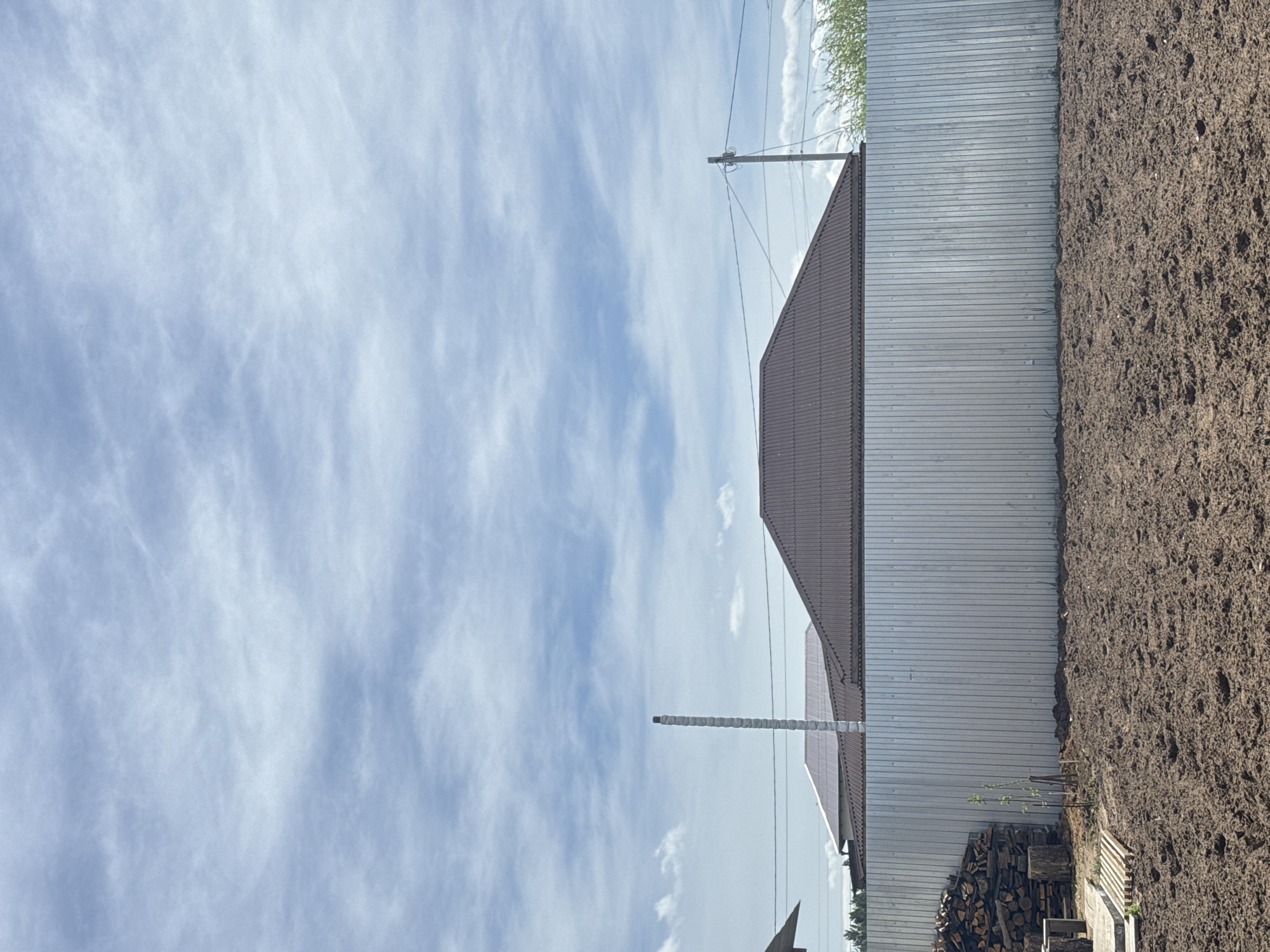 The image shows a landscape with a mix of natural and man-made elements. The sky is filled with wispy clouds. A building with a brown roof is partially visible. A corrugated metal fence runs alongside a patch of tilled soil. Firewood is stacked near the fence. Power lines are also visible against the sky. The location is Unknown, Unknown.