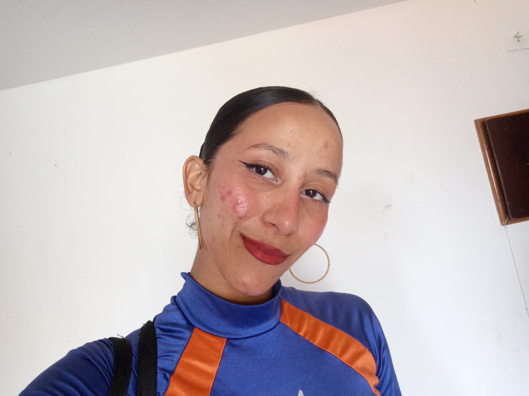 A young woman poses for a selfie indoors against a plain white wall. She has dark hair pulled back, eyeliner, and lipstick. She wears a blue long-sleeved shirt with orange stripes on the shoulders. She is wearing gold hoop earrings. A portion of a black strap of a bag is visible on her left shoulder. There is no indication of Porlamar, Venezuela, or any other specific location.