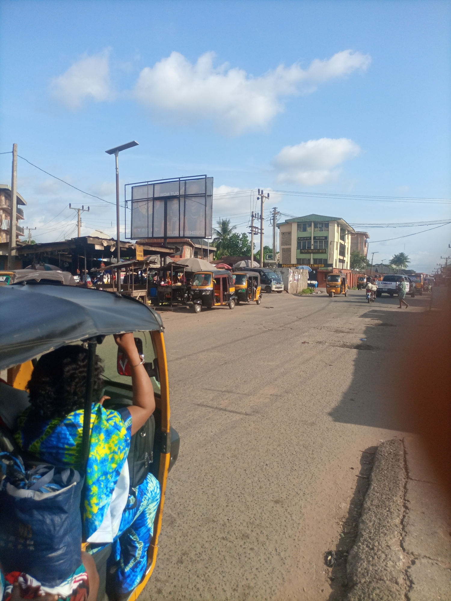 This media file captures a street scene in Nkpor, Nigeria, likely during the daytime. The shot is taken from the perspective of someone riding in a yellow auto-rickshaw, with the passenger's colorful blue and yellow patterned clothing visible in the foreground. The auto-rickshaw itself is a prominent object in the scene, with its dark canopy and yellow exterior.
The street is paved with asphalt, showing signs of wear and tear, and is bustling with activity. Several other auto-rickshaws are present, along with other vehicles including a white pickup truck and a few cars. A motorbike is also visible with a rider.
Along the sides of the street are various stalls and small shops, some covered with corrugated iron roofs and awnings, suggesting a market or commercial area. A large billboard is mounted on a metal frame, with some text partially obscured, but "OUT NOW" is legible on it. A multi-story building with a green roof and the text "PACIFIC HOUSE" and "PACIFIC PUBLISHERS LIMITED" is a notable landmark in the background. Power lines and utility poles crisscross the sky, typical of urban or semi-urban settings.
The sky is a bright blue with scattered white clouds, indicating fair weather. The lighting suggests it is either late morning or early afternoon. The overall atmosphere appears to be one of everyday commerce and transportation, with people engaged in their daily routines. The scene depicts a vibrant and active street in a Nigerian town.