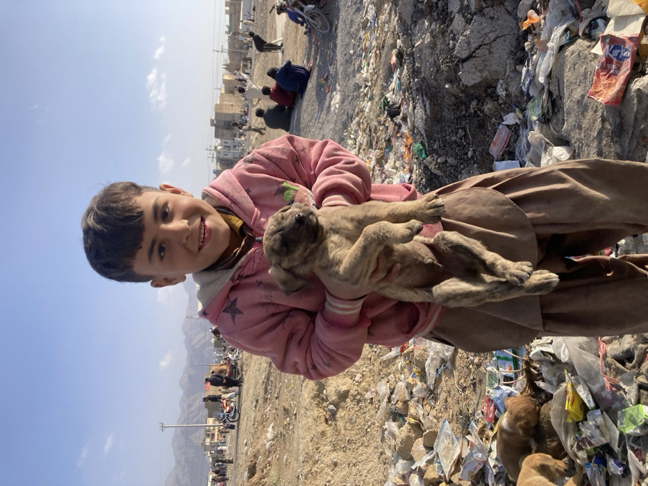 A young boy, approximately 8-12 years old, stands centrally in the foreground of an outdoor scene in Quetta City Tehsil, Pakistan. He has short dark hair, dark eyes, and is smiling directly at the camera, revealing his teeth. He wears a worn and stained pink hooded sweatshirt over a visible yellow undershirt, and loose-fitting light brown trousers. He holds a brindle-patterned puppy in his arms, which appears to have its eyes closed or partially closed and its paws hanging downwards. The immediate ground around the boy is covered with dirt, rocks, and an accumulation of refuse, including plastic bags, wrappers, and assorted trash. To the boy's lower left, another light brown puppy is visible amidst the debris.
The background shows a vast, open, unpaved area with numerous individuals scattered across the mid-ground, some standing, some seated on the ground. Several motorcycles are visible. Further back, modest buildings and utility poles with overhead wires are present. The distant horizon is dominated by arid, rocky mountains under a clear, pale blue sky with scattered white clouds. The overall environment suggests a market or a populated, undeveloped area.