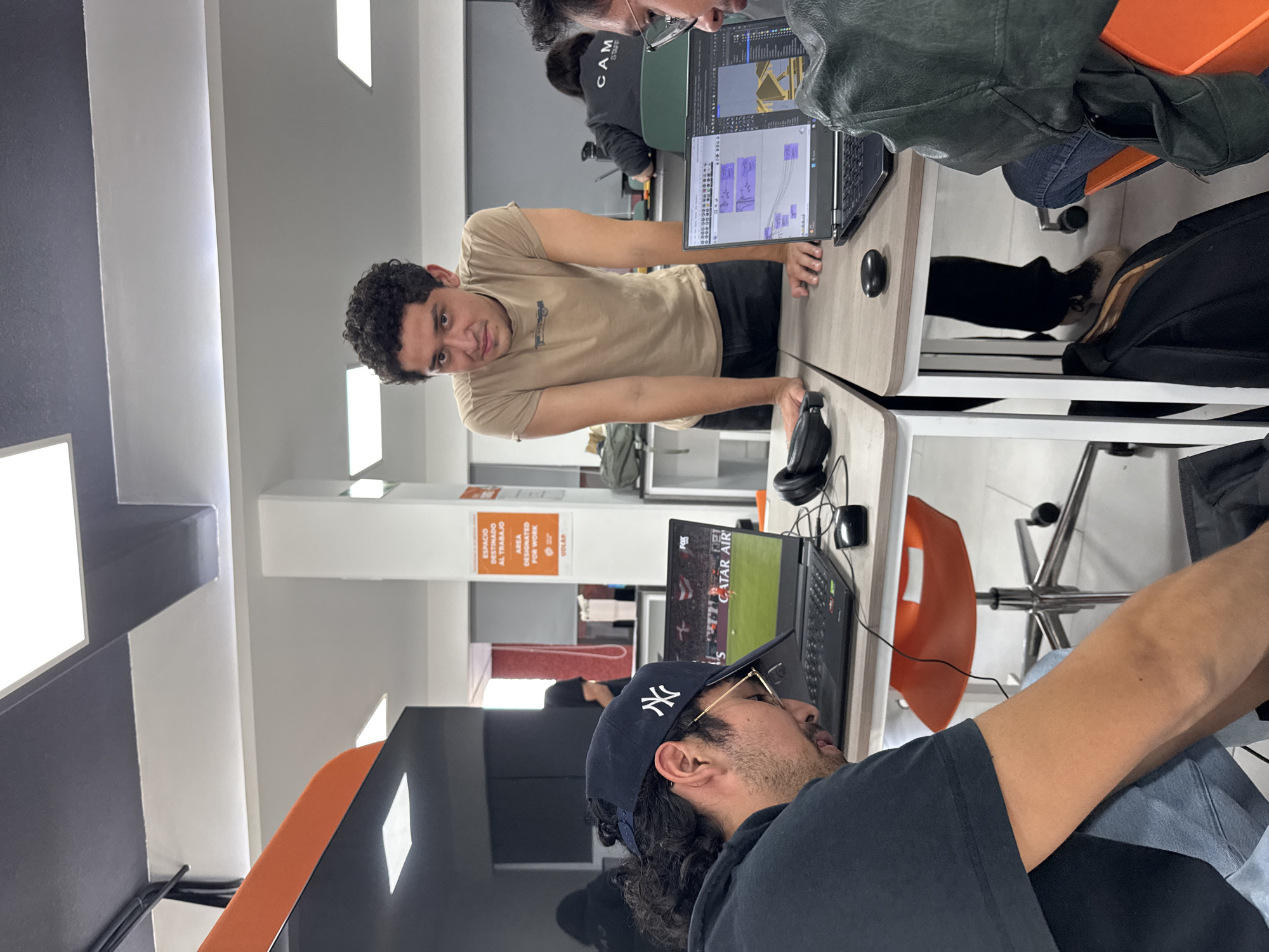 The photograph shows an indoor scene, possibly an office or study area in San Andrés Cholula, Mexico. Three individuals are present, engaged in activities involving laptops.
One man in a tan t-shirt is leaning across a table and looking towards the camera. He has dark, curly hair and is wearing a casual t-shirt. Another individual, partially visible, is working on a laptop. Another person wears a baseball cap with a logo on it, along with glasses, and seems to be watching a soccer game on a laptop.
The laptops are on tables, and there are office chairs around them. A sign is visible on the wall with the text "ESPACIO DESTINADO AL TRABAJO AREA DESIGNATED FOR WORK UDLAP" written on it. The environment appears to be a workspace with a modern design.