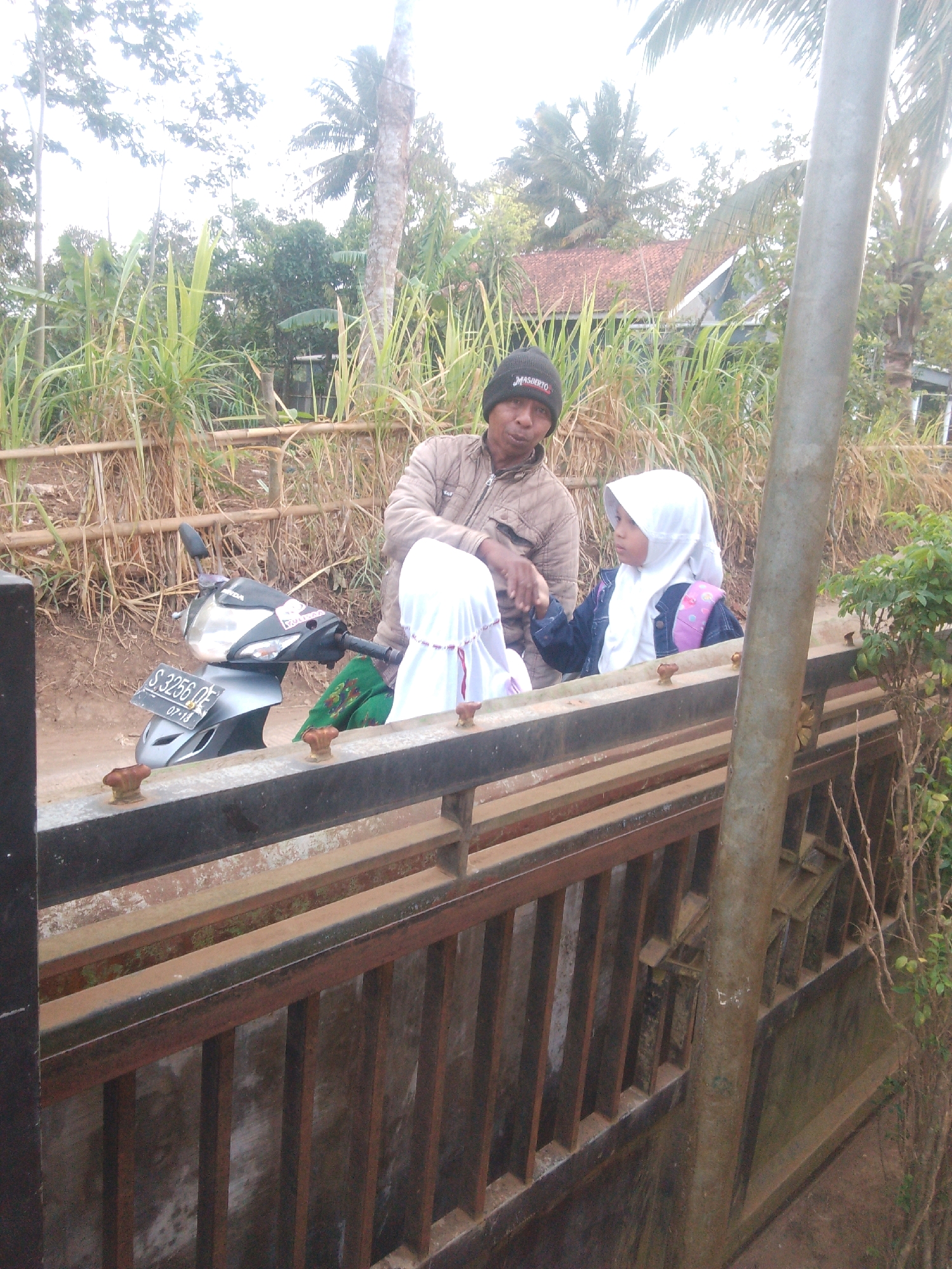 In Betek, Indonesia, a man wearing a black beanie and a light brown jacket stands next to a motorcycle. He is looking towards the camera. Beside him, two children are also present. One child wears a white hijab and a dark blue jacket, and the other child also wears a white hijab. The children appear to be looking away from the camera. They are positioned behind a metal fence. The background consists of tall green plants, trees, and a house with a terracotta roof. The ground is covered in dirt. The scene appears to be outdoors in a rural or semi-rural setting.