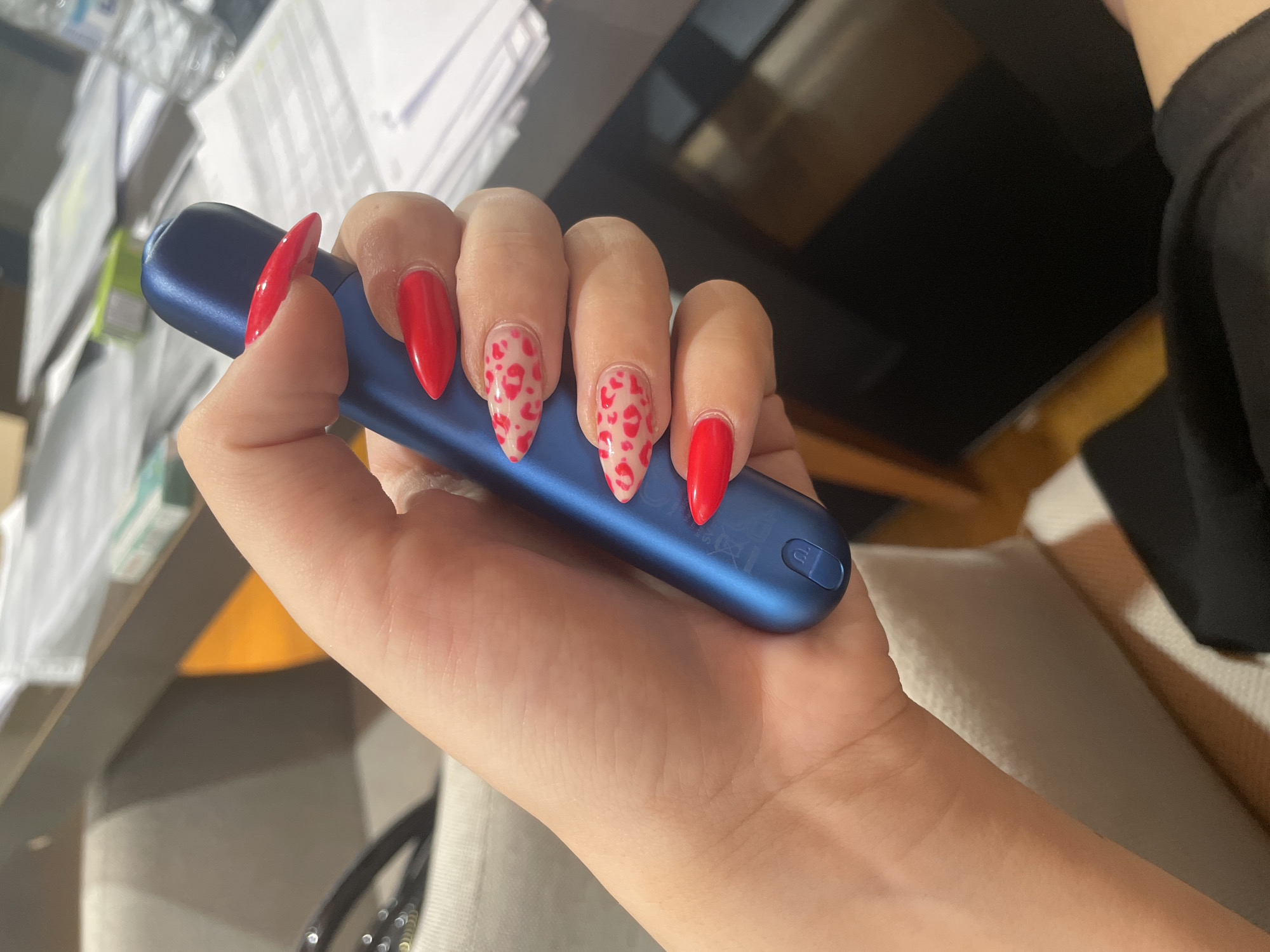 A close-up, overhead shot shows a person's hand holding a blue electronic cigarette device. The hand has long, stiletto-shaped fingernails painted in bright red, with some nails featuring a leopard print design of red hearts on a light pink background. The background appears to be an indoor setting with a desk covered in papers and office supplies, including a bottle of water. The lighting is bright, suggesting an office or study environment. The location in Burgas, Bulgaria, cannot be determined from the visual information in the image.