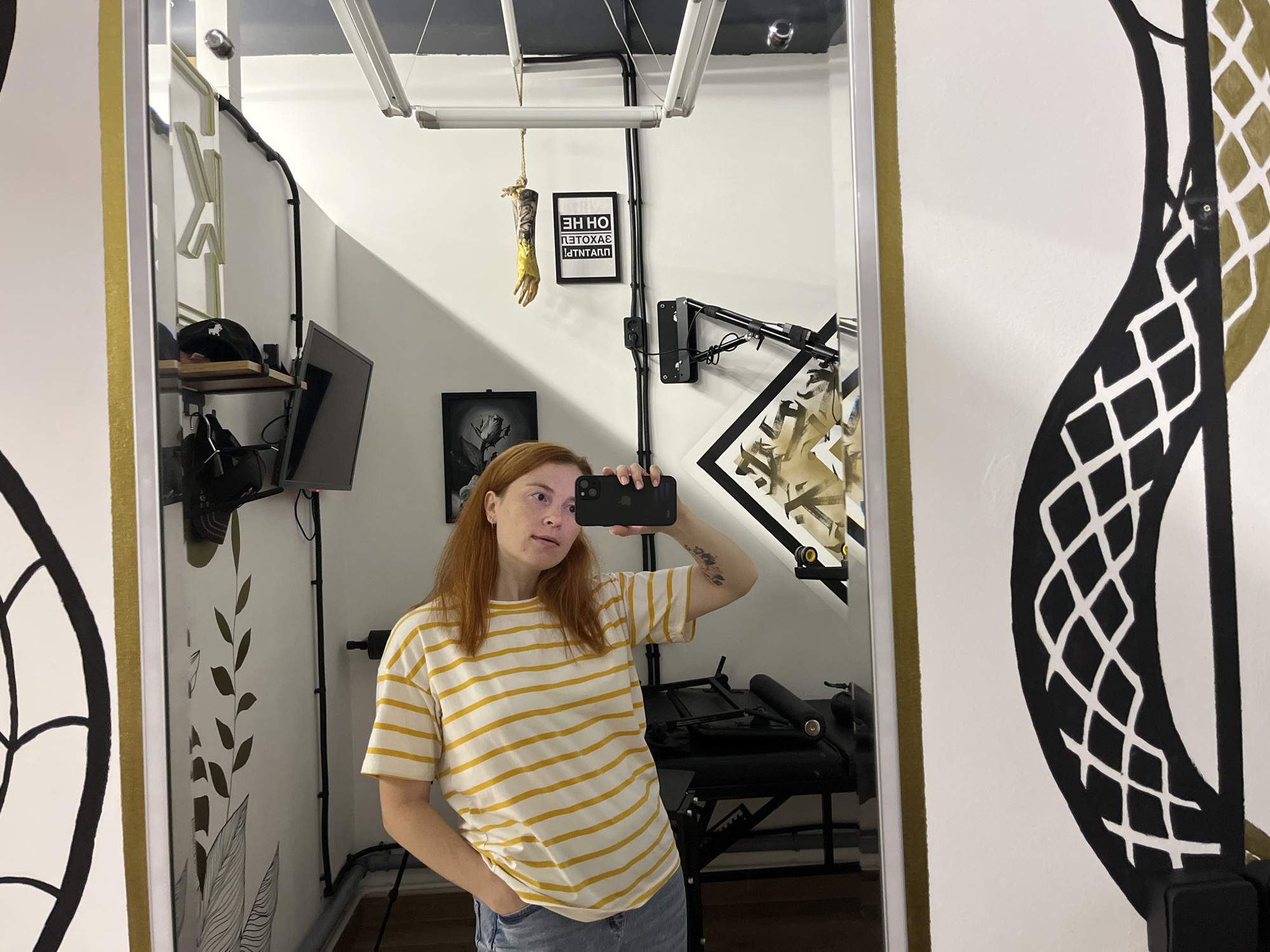 A person with reddish-brown hair, wearing a yellow and white horizontally striped t-shirt and blue jeans, stands in front of a mirror and takes a selfie. The person's left hand is in their pocket, and their right hand is holding a black smartphone up to take the photo. The background reflected in the mirror shows a room with white walls. On the wall, there are several objects including a framed picture of a rose, a framed sign with text that appears to be backwards, and a detached prosthetic arm hanging from a rope. There are also shelves with items on them and various black electrical conduits and fixtures. A large decorative black and gold graphic design is visible on the right side of the image, partially obscuring the mirror. The lighting appears to be from fluorescent fixtures on the ceiling. The overall environment suggests an artistic or studio space. This image is not located in Chaikovsky, Russia.