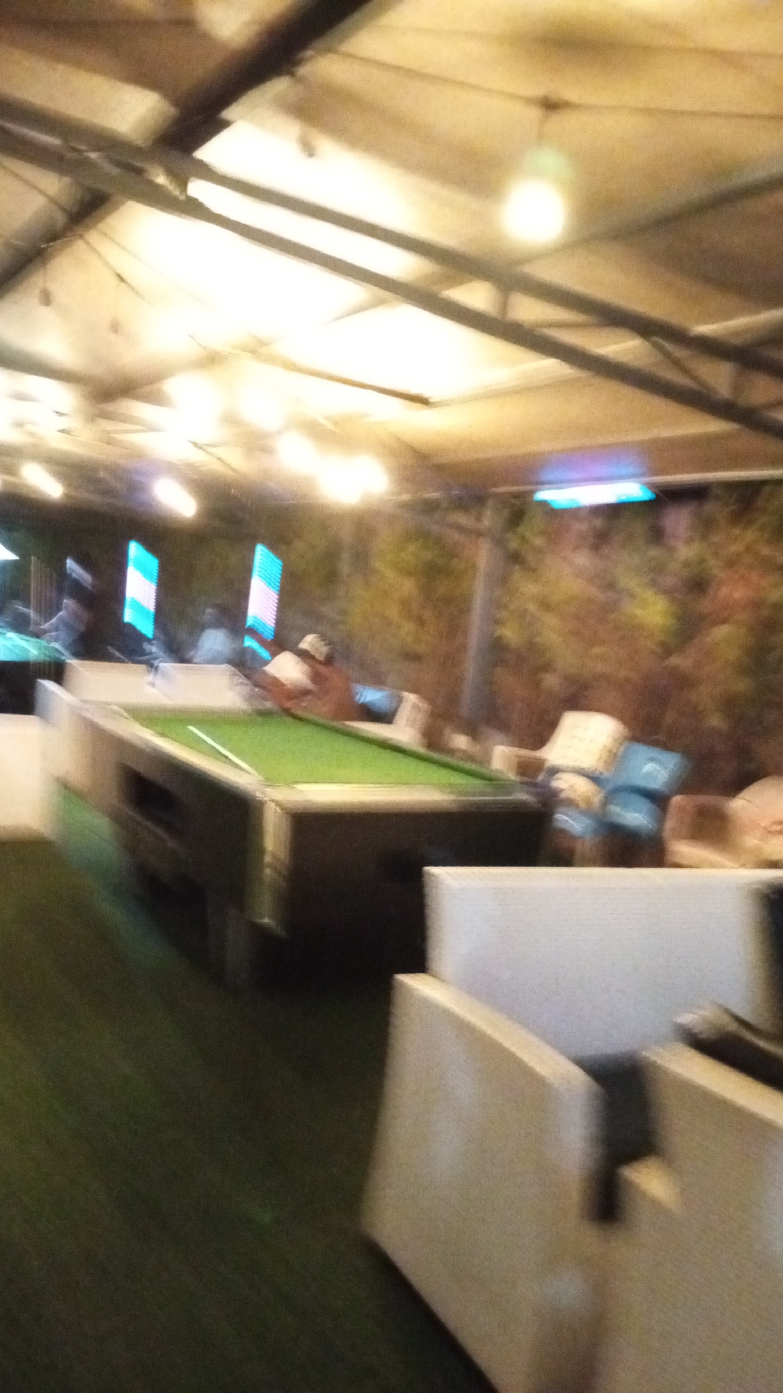 The image depicts an indoor recreational area in Adankolo, Nigeria, with a pool table as the central focus. A person is leaning over the pool table, which is covered in green felt. White and beige seating is arranged around the table. The floor is covered in artificial green turf. The building has a metal roof structure. Interior lighting is prominent. In the background there are vertical rectangular lighting panels. Outdoor elements can be seen, suggesting the building has open sides. The image has motion blur.
