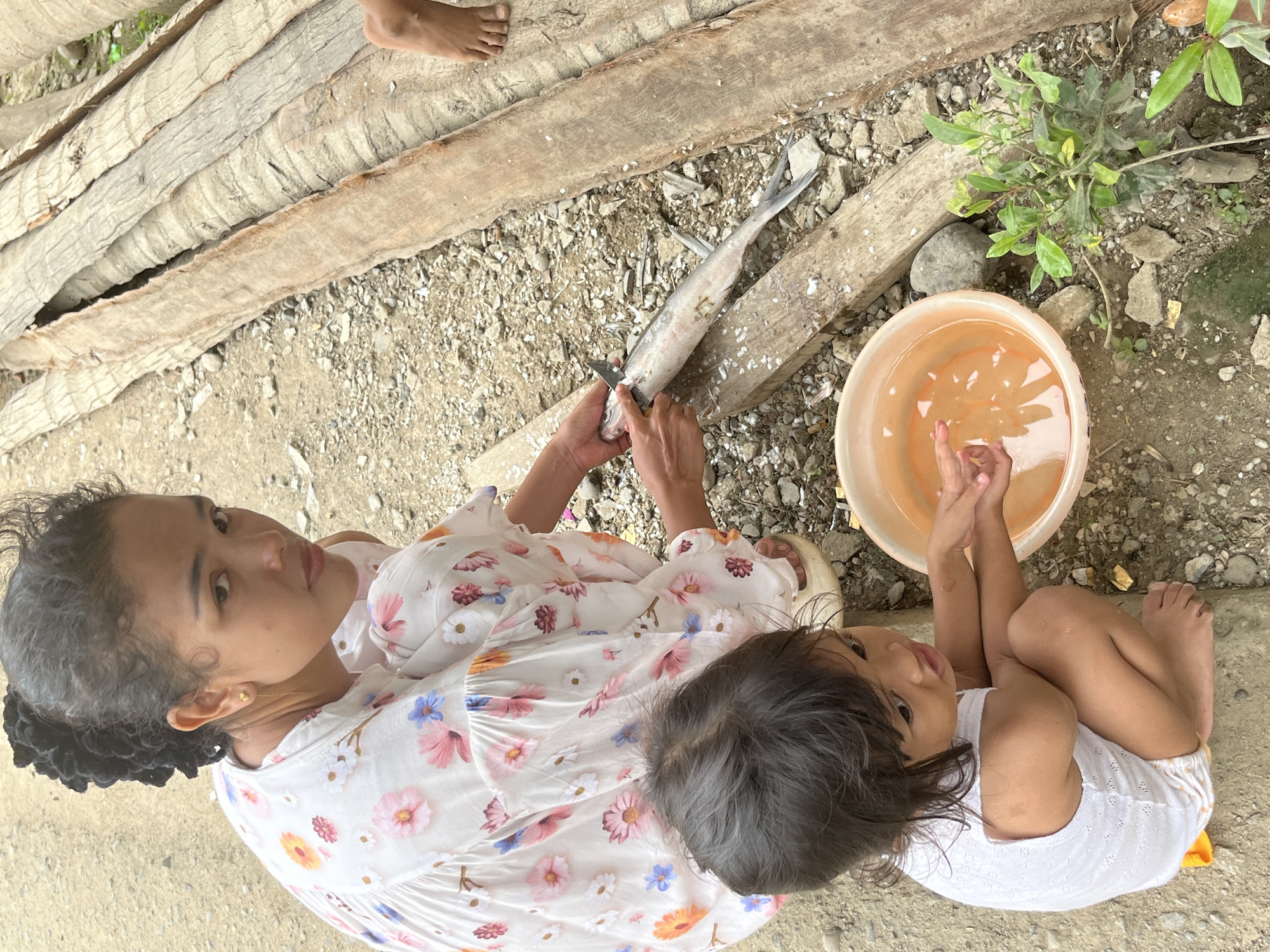 In Kruengmate, Indonesia, a woman in a floral dress is cleaning a fish on a wooden surface. Beside her, a young child in a white outfit crouches near a plastic bowl filled with orange water. The ground is covered in gravel and small stones, with a few green plants growing nearby. In the background, large wooden logs are stacked.
