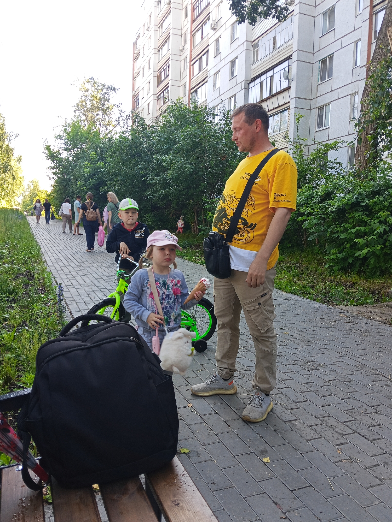 A man in a yellow t-shirt and tan cargo pants stands on a brick pathway next to a building with a light-colored facade. He is looking down and to his left. In front of him, a young girl in a pink hat and a gray shirt with a flamingo print is holding an ice cream cone and a fluffy white toy. A small green bicycle is positioned between her and a young boy in a black jacket and green-and-white baseball cap. Several other people are walking on the pathway in the background, and dense green trees line the side of the path. A large black backpack rests on a wooden bench in the foreground. The scene appears to be a public park or walkway in Chaikovsky, Russia, given the architectural style of the building and the natural setting.