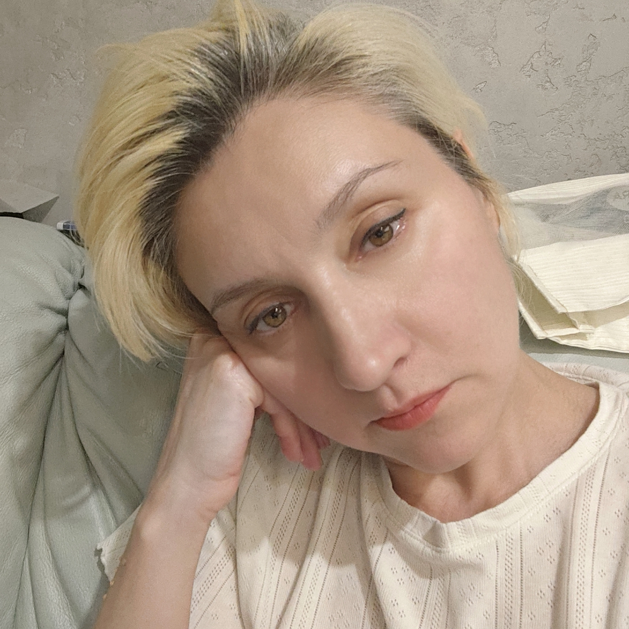 The image shows a close-up of a woman's face. She has short, blonde hair with dark roots, indicating it is dyed. Her eyes are brown, and she is wearing eyeliner. Her lips are painted a peachy-pink color. She is resting her head on her hand, and her expression appears thoughtful or somber. The background is slightly out of focus, showing a textured gray wall and what appears to be a light-colored couch or upholstered furniture. There is also a folded light-colored fabric visible behind her. No specific details about Novosibirsk, Russia are present in the image.