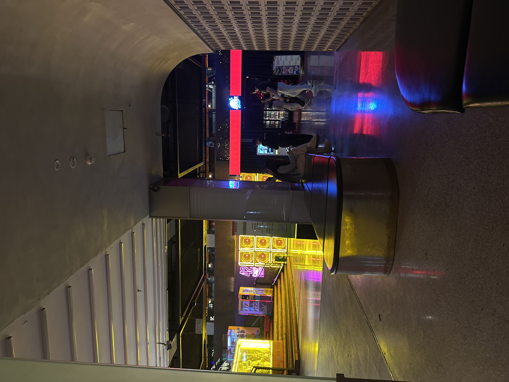 Here's a matter-of-fact description of the scene:
The image depicts an interior space in the Liangjiang area of Chongqing, China, characterized by modern architectural features and bright, multicolored lighting. People are present in the distance, some seated and others standing around a bar or counter area. A cylindrical structure is prominent in the foreground, potentially serving as seating or a decorative element. Illuminated advertisements and signage are visible within the complex, adding to the lively ambiance of the location. The architecture includes archways, structured paneling, and various decorative lighting fixtures.