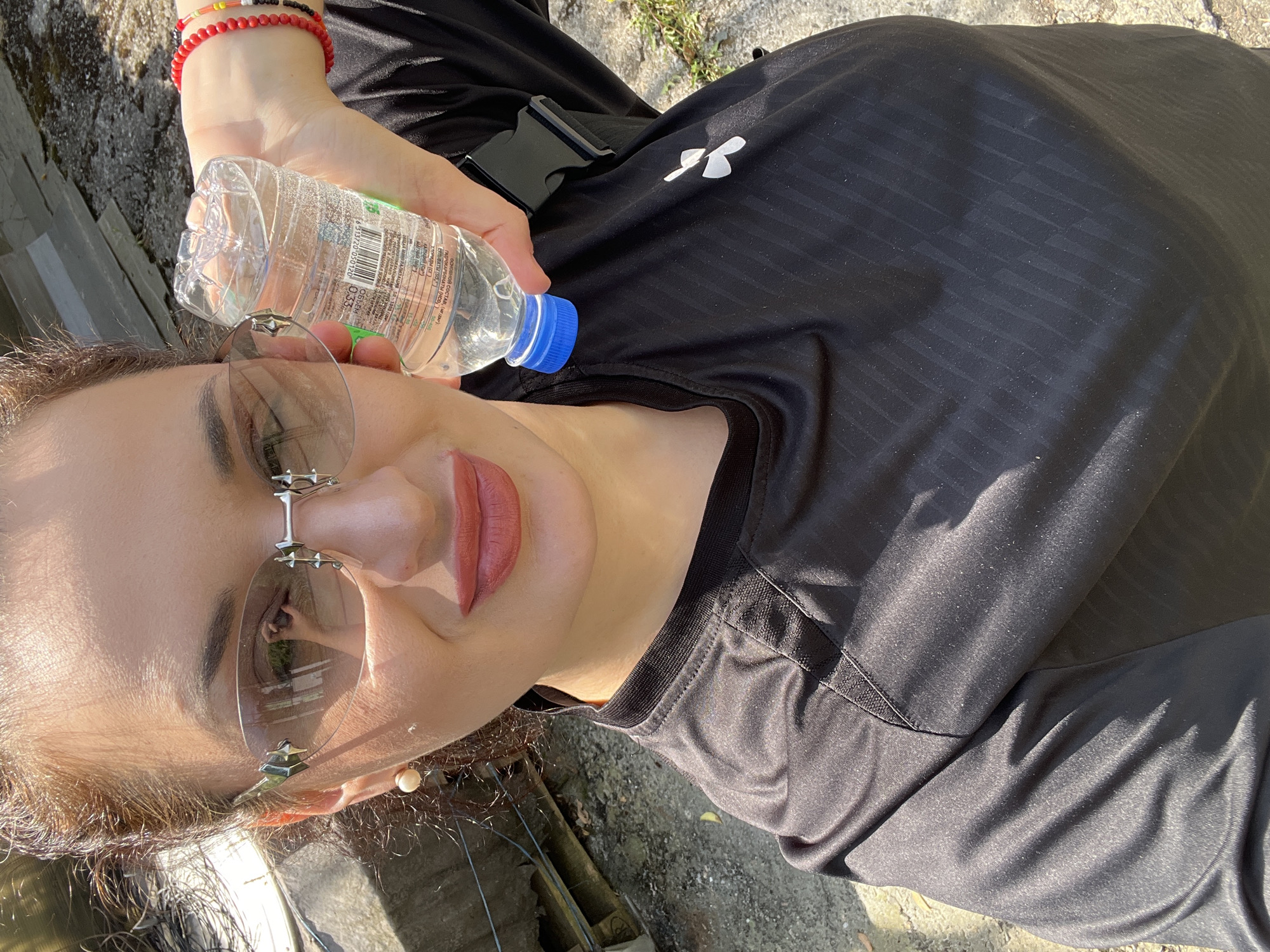 A selfie captures a woman from the chest up, outdoors under bright sunlight in the city of Unknown, Unknown. She has dark hair pulled back and fair skin, with a faint smile and reddish-brown lipstick. Her right ear features a pearl stud earring. She wears rimless sunglasses with light-tinted lenses and decorative silver/grey frames; the left lens reflects a hand holding a phone, indicating a selfie. She is dressed in a black, short-sleeved athletic top with a subtle textured pattern of diagonal stripes and a white stylized 'U' or inverted 'A' logo on the left chest. A black strap with a plastic buckle, possibly from a waist pack, is visible on her right side. Her left wrist displays multiple beaded bracelets, predominantly red, and another with red, yellow, and black beads. She holds a clear plastic water bottle with a blue cap in her left hand, positioned near her head; the bottle contains clear liquid and its label shows "0.53 L" and Cyrillic text. The background features an unpaved or concrete ground with sparse vegetation and what appear to be remnants of structures or debris. Strong shadows are cast across her face and shirt, consistent with direct sunlight.