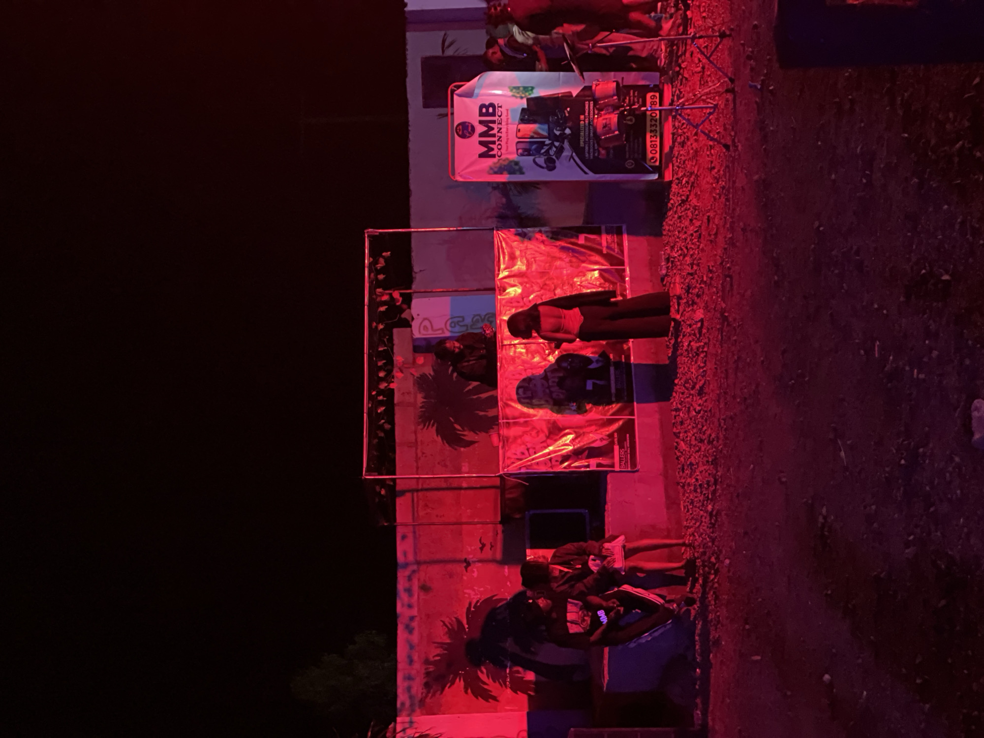 The image depicts an outdoor nighttime scene illuminated by vibrant red lighting, potentially indicating an event or gathering in Durumi, Nigeria. In the background, a wall is painted with tropical motifs of palm trees. A makeshift stage or platform is set up, adorned with red translucent fabric and displaying graphics, including the "MMB CONNECT" logo with images of mobile phones and headphones. A banner for "MMB CONNECT" is also prominently displayed to the right, listing services like "SPECIALIZED IN LAPTOPS, SMART PHONES, MACBOOKS | SMART ACCESSORIES." A phone number, "0813332089," is visible on the banner.
Several individuals are present. On the left, two people are seated on a raised blue structure. To the right of the stage, a person in a white top and black pants stands facing away from the camera, seemingly engaged with the event. Further to the right, a group of people are gathered, some appearing to be performers or attendees. Musical equipment, including what looks like a drum set and microphone stands, is visible on the right side of the scene. The ground in the foreground is covered with gravel and debris. The overall atmosphere suggests a lively event possibly related to technology or entertainment.