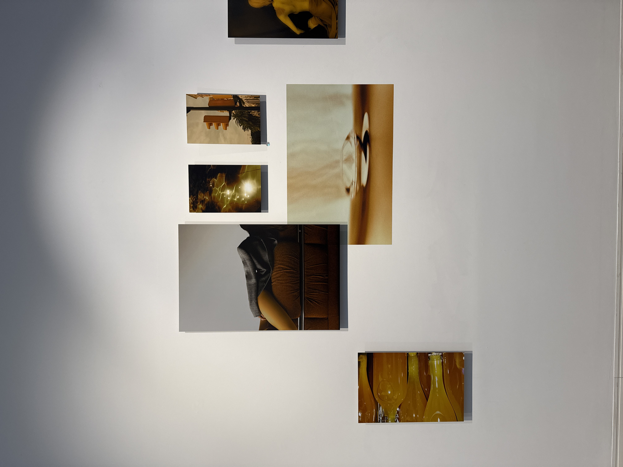 The image shows a white wall on which several photographic prints are displayed. The prints depict various subjects: a classical sculpture, a traffic light, a scene with illuminated reflections in water, an abstract image focusing on a doorknob or handle, a person in a partial suit, and a collection of yellow bottles. The context of the images suggests an art exhibition or gallery setting. The exact location in Sofia, Bulgaria, is not discernible from the visual information provided.