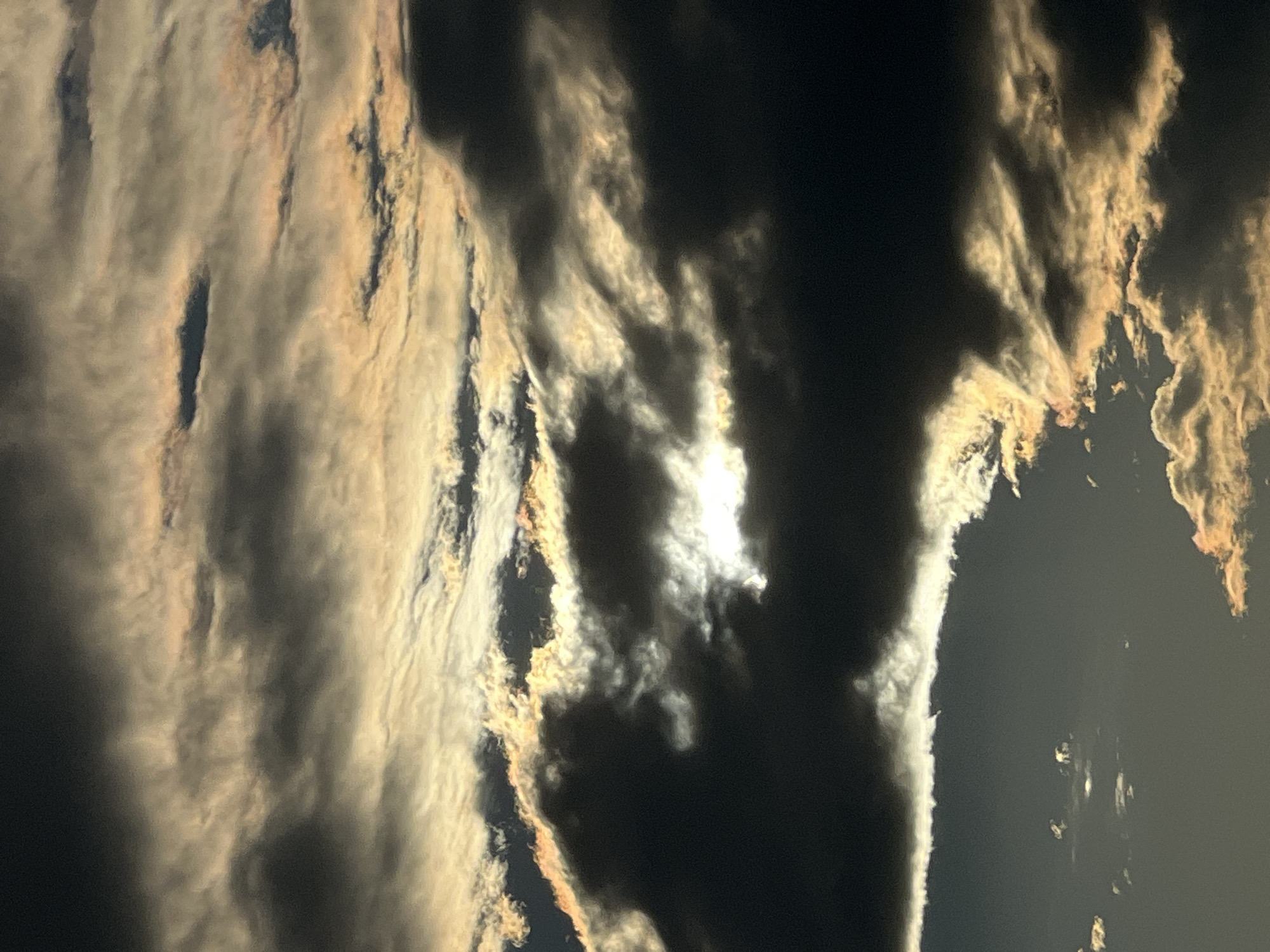 The image shows a sky filled with dramatic clouds illuminated by the sun. The clouds are dark and heavy in some areas, while others are thin and wispy, catching the light with hues of orange and yellow. The sun is partially obscured by the clouds, creating a bright, ethereal glow. The overall scene is one of a dynamic and possibly stormy sky. There are no people, subjects, objects, or actions depicted in this image, nor is there any indication of a specific location within the city of Saint Petersburg, Russia.