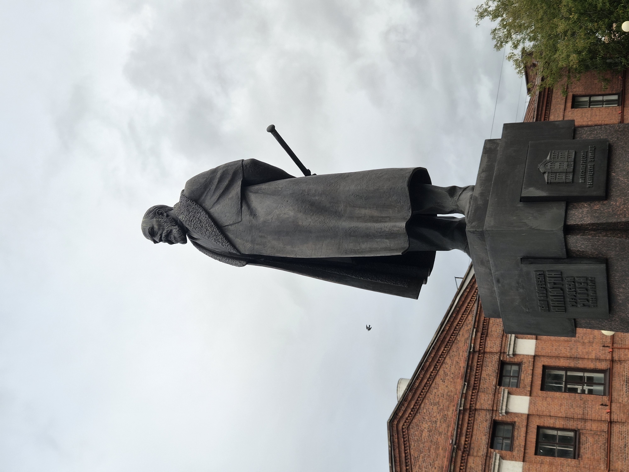 The image shows a monument to Pavel Andreyevich Florensky located in Cherepovets, Russia. The statue depicts a man with a beard, wearing a long coat with a fur collar, and holding a walking stick behind his back. He stands on a large, dark stone pedestal. Inscriptions and an emblem of a building are visible on the pedestal. To the left of the statue is a red brick building with multiple windows. A bird is captured in flight in the overcast sky. The overall scene suggests an outdoor, public space in a city.