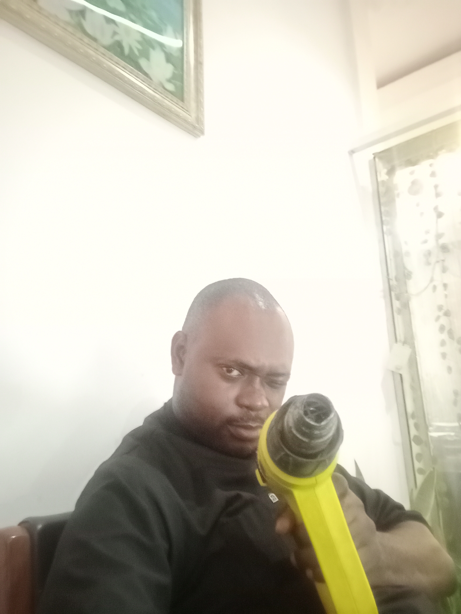 A bald, dark-skinned man in a black shirt holds a yellow heat gun towards the camera with a focused, squinting gaze. He is seated. A framed picture hangs on a white wall in the background, with a window to the right. The location is presumably an interior space in Gwarinpa, Nigeria, though this is not explicitly shown in the image.