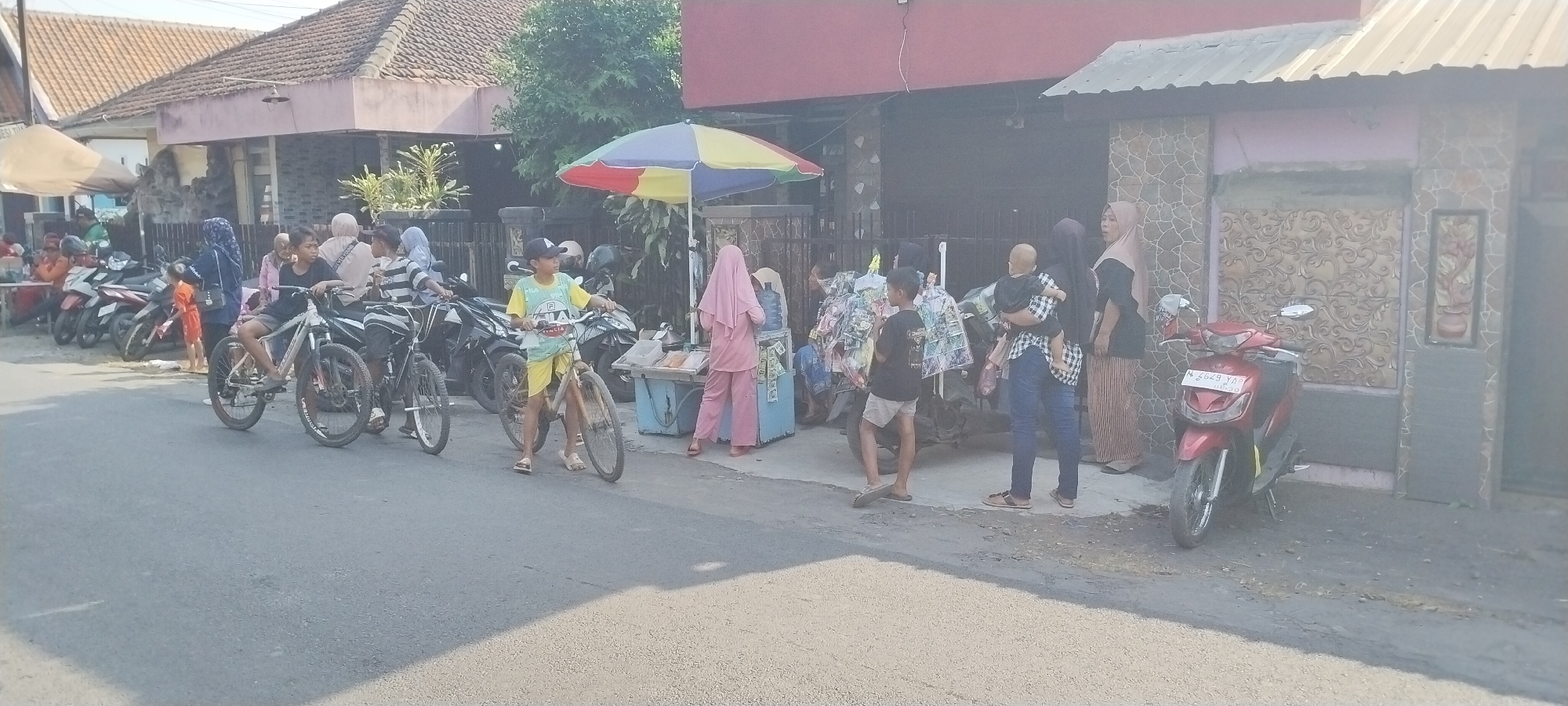 The image depicts a street scene in Yosowilangun Kidul, Indonesia. A group of people, including several children and adults, are gathered along the side of a paved road. Several motorcycles are parked on the side of the road, and two bicycles are visible, with one being ridden by a child. A woman in a pink hijab and matching outfit stands at a small stall selling drinks and snacks, which is covered by a colorful umbrella. Another child in a yellow t-shirt and shorts stands beside a bicycle. In the background, there are buildings with tiled roofs and textured walls. A red scooter is parked in front of one of the buildings. The overall atmosphere appears to be that of a typical street vendor and community gathering on a sunny day.