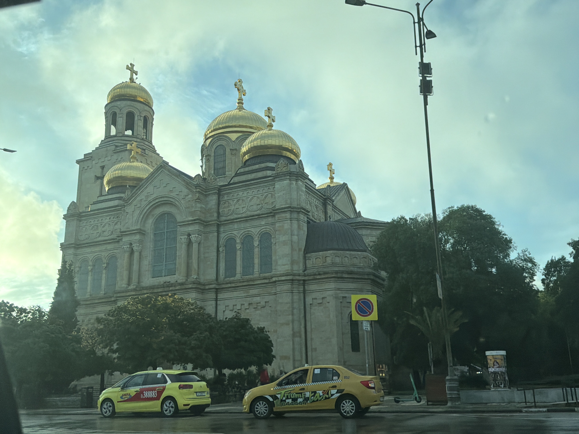 The image displays a grand Eastern Orthodox cathedral featuring multiple golden domes topped with crosses, set against a cloudy, bright sky. The cathedral is constructed from light-colored stone, adorned with arched windows and intricate architectural details.
In the foreground, a wet street reflects the surrounding elements, suggesting recent rain. Two yellow taxis are visible; the one on the left is a hatchback with "MEGA TAXI" and the number "388883" printed on its side in Cyrillic script. The taxi on the right is a sedan featuring "Triume! Taxi" and the number "644444", also in Cyrillic. Lush green trees and bushes line the street, partially obscuring the lower portions of the cathedral.
Street furniture includes a tall street lamp post on the right, a traffic sign indicating "no parking" or "no stopping," and an electric scooter parked near the curb. A faint figure in red clothing can be seen near the left taxi, partially hidden by trees. The scene takes place during the daytime under an overcast sky, providing diffused lighting. The combination of the distinctive church architecture and the Cyrillic text on the taxis strongly indicates a location in Eastern Europe, likely Bulgaria, with the building bearing a resemblance to the Dormition of the Theotokos Cathedral in Varna.