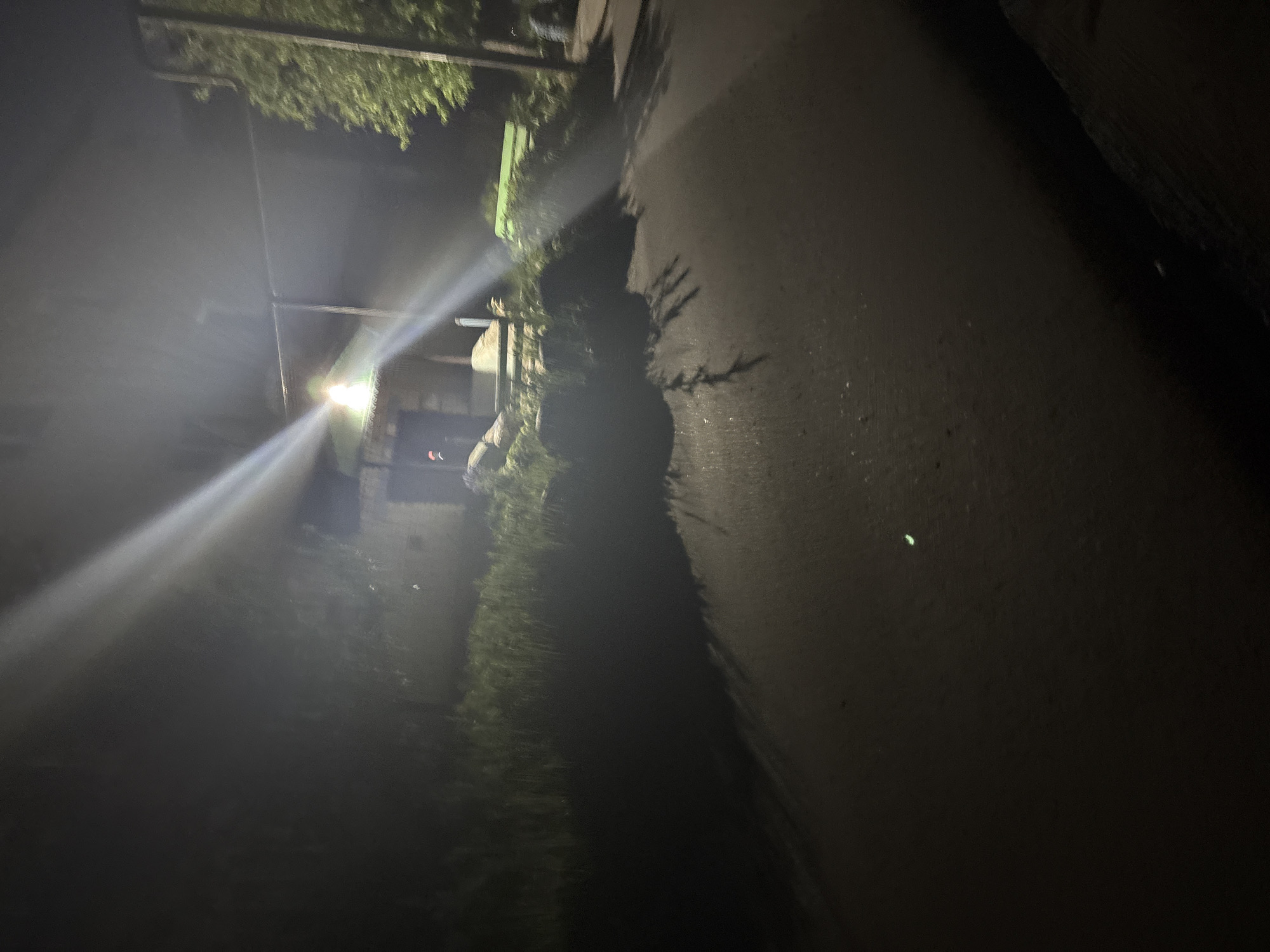 The image displays an outdoor nocturnal scene, primarily illuminated by a strong artificial light source positioned in the upper-center of the frame, which produces significant glare and distinct light rays. A dark, paved surface, resembling a path or road, extends from the foreground into the mid-ground, showing texture and reflections. Elongated shadows from unseen objects are cast across the left side of this surface. A small, bright green spot is visible on the path in the lower-left quadrant. Dense, dark vegetation, including trees with visible green foliage, flanks both sides of the path. In the mid-ground, to the right, a green bench is partially discernible among the foliage. A building with a dark structure and a lighter roofline is situated in the mid-ground, illuminated by the central light source. No individuals are present in the scene.