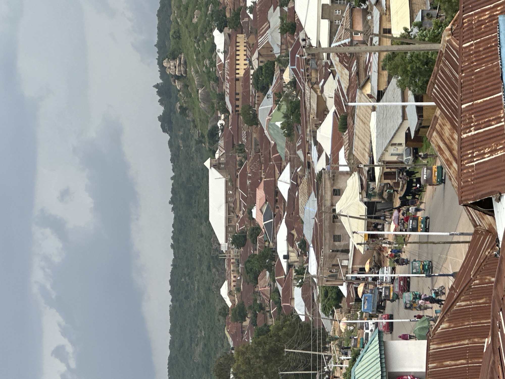 This is an aerial view of a city street in Jos, Nigeria, with buildings and a hilly landscape in the background. The foreground shows several buildings with corrugated metal roofs, some of which are rusted and weathered. A paved road runs through the scene, with several vehicles including cars, a truck, and auto-rickshaws. People are visible walking on the sidewalks and near the vehicles, some under umbrellas. The middle ground consists of a dense collection of houses and buildings, many with similar corrugated roofs and some with white or grey modern roofs. A large building with a white roof and a church-like steeple is prominent in the upper middle section. The background features a verdant, hilly terrain with scattered rock formations and trees, under a cloudy sky. The overall impression is of a bustling urban environment nestled within a natural landscape.