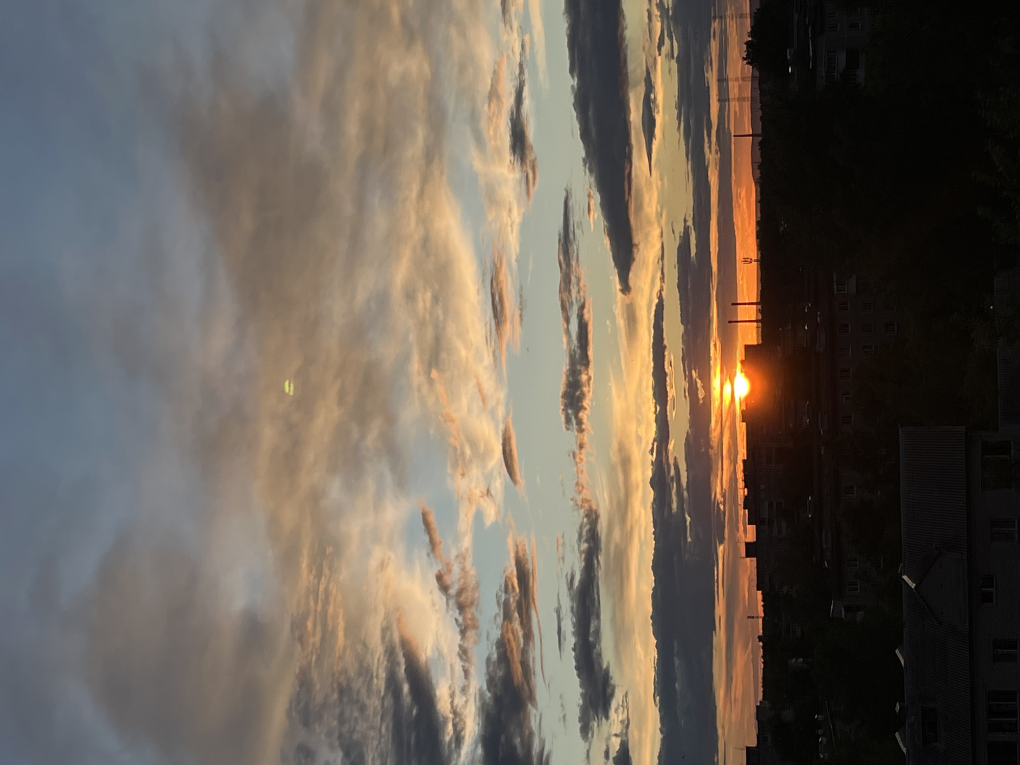 The image shows a sunset over an urban landscape in an unknown city. The sun is low on the horizon, appearing as a bright orange orb. The sky is filled with clouds that are illuminated by the setting sun, displaying a range of colors from light pink to dark grey. Below the horizon line, the silhouettes of buildings and industrial structures are visible against the bright sky. The scene is characterized by the dramatic contrast between the bright, colorful sky and the dark, silhouetted buildings.