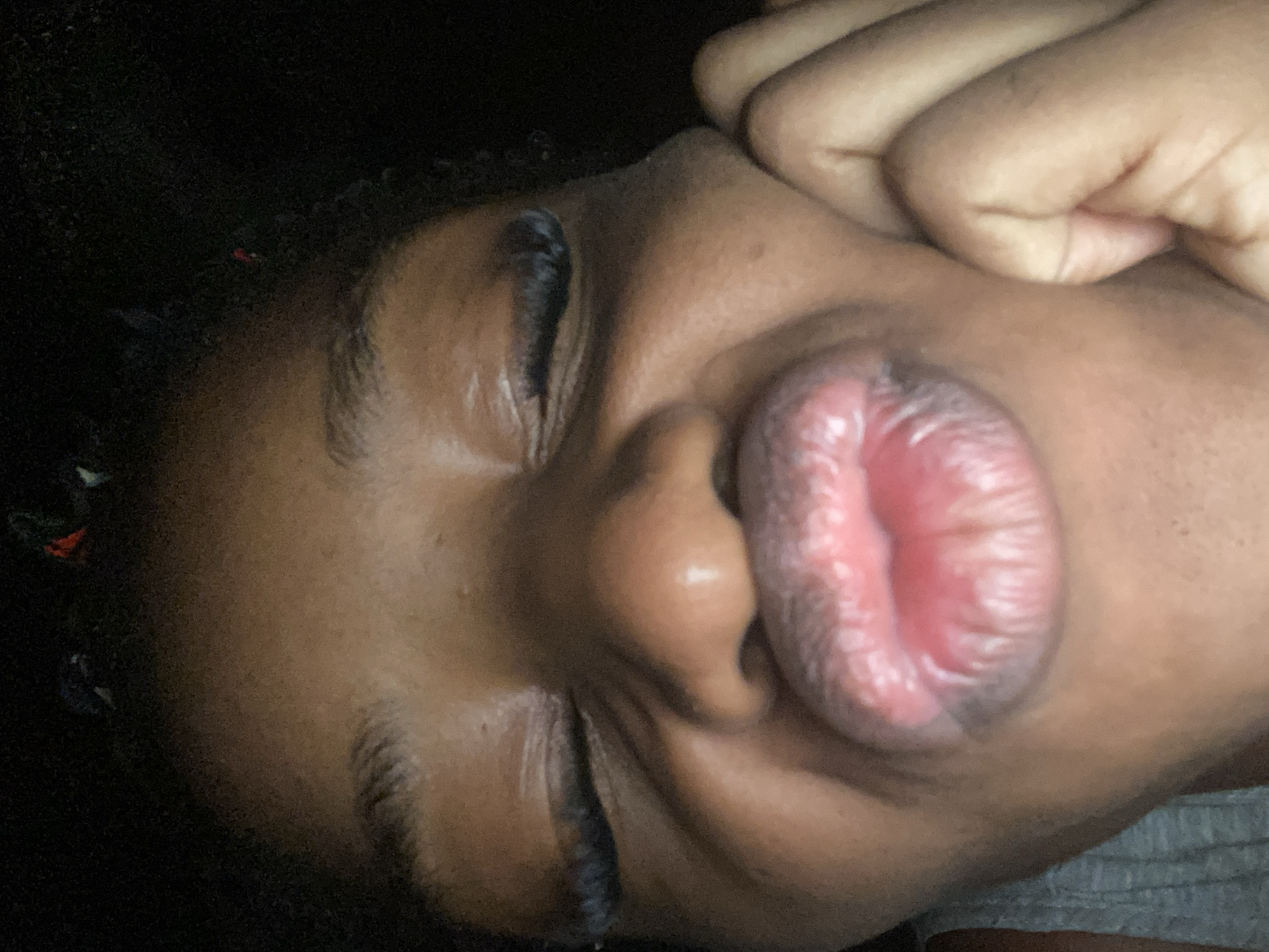 The image is a close-up, low-light, selfie-style photograph of a person's face, featuring prominent lips pursed in a kiss gesture. The individual's eyes are closed, and long eyelashes are visible. Their skin tone is dark. The background is dark and indistinct, suggesting an indoor setting or nighttime. A hand is visible on the right side of the frame, resting near the person's cheek. The image does not contain any identifiable landmarks, street scenes, or architectural features that would allow for its localization to Jos, Nigeria, or any specific urban environment.