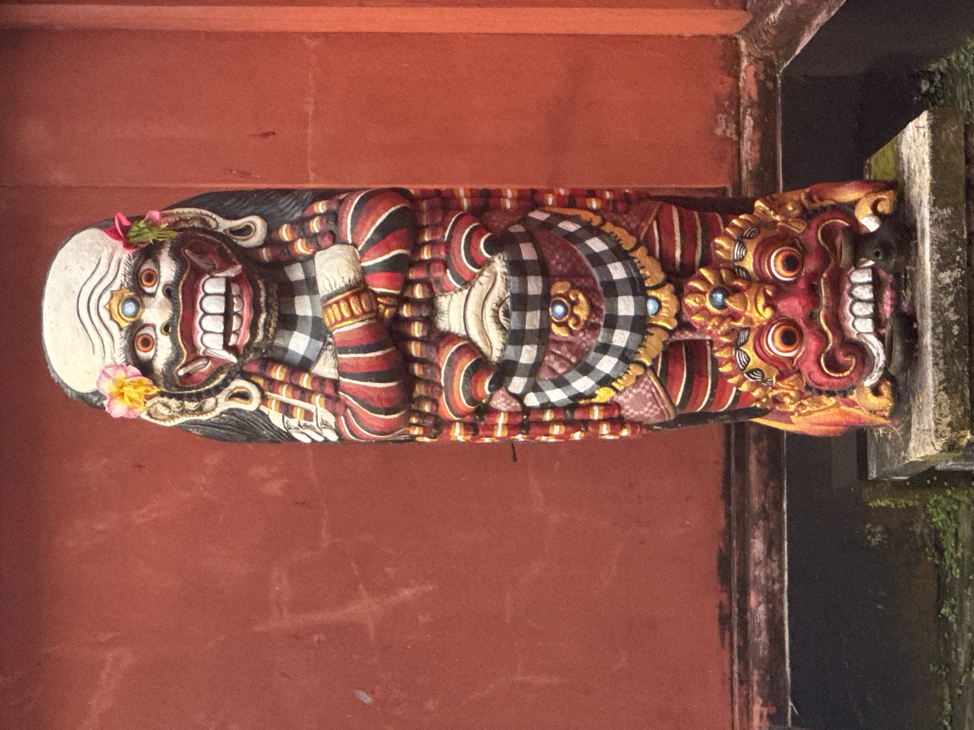 The image shows a richly decorated, horizontal wood carving of three grotesque figures, mounted on a red-painted wall and part of a wooden post. The carving appears to be part of a traditional Balinese architectural element, likely found in Denpasar, Indonesia. From left to right, the first figure has a white face with incised lines, wide eyes, a black beard, and a large, toothy grin. A small pink flower adorns its head. The middle figure is wrapped in black and white checkered cloth and has a blue stone set in its forehead. The third figure is predominantly red and gold with large, circular eyes and a fierce expression. A portion of a weathered, moss-covered wooden post is visible to the right of the carving.
