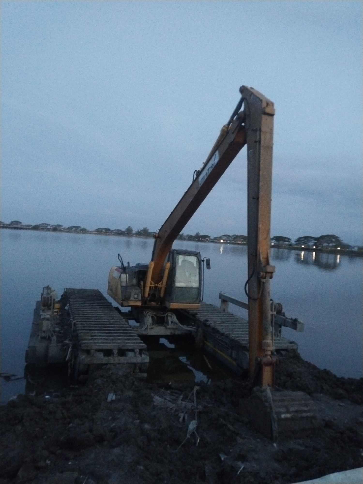 An amphibious excavator stands partially submerged in water near a muddy bank in Lhokseumawe, Indonesia. The yellow machine is equipped with tracks for traversing both land and water. The excavator arm is raised, with its bucket resting on a mound of dark mud. The water stretches into the distance, reflecting lights from buildings or structures on the opposite shore. The sky is overcast, suggesting dusk or dawn. Trees are visible on the shoreline in the distance.