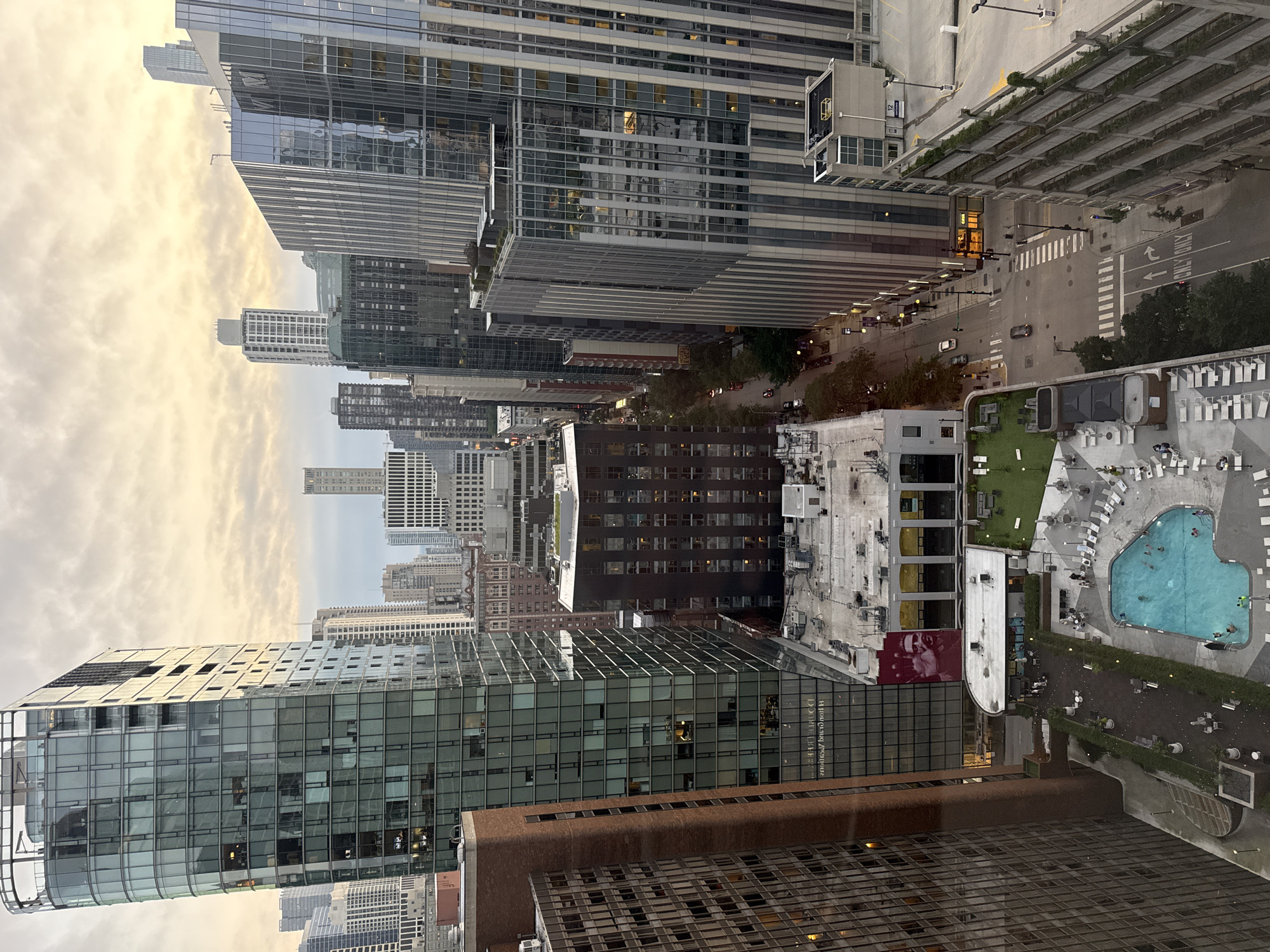 This image captures an aerial view of a dense urban environment, likely Chicago, given the architectural style and the presence of a prominent swimming pool area. The scene is dominated by numerous high-rise buildings constructed with glass and steel, reflecting the cloudy, overcast sky which is illuminated with warm, golden light, suggesting it might be dawn or dusk.
In the foreground, a large, irregularly shaped swimming pool is filled with people swimming and lounging. Surrounding the pool are lounge chairs and a patio area with tables and umbrellas. Adjacent to the pool area is a green space, possibly a rooftop garden, with seating. Several low-rise buildings, likely part of a hotel or residential complex, are visible around the pool and garden.
A street runs through the scene, with cars moving and parked. The street is lined with trees and streetlights, and crosswalks are clearly marked. Various buildings of different heights and architectural designs are densely packed, creating a classic cityscape. Some buildings have visible signage, though the text is not entirely legible. A large mural of a person's face is visible on the side of one building. The overall impression is of a bustling, modern city center with amenities for recreation and residential living.