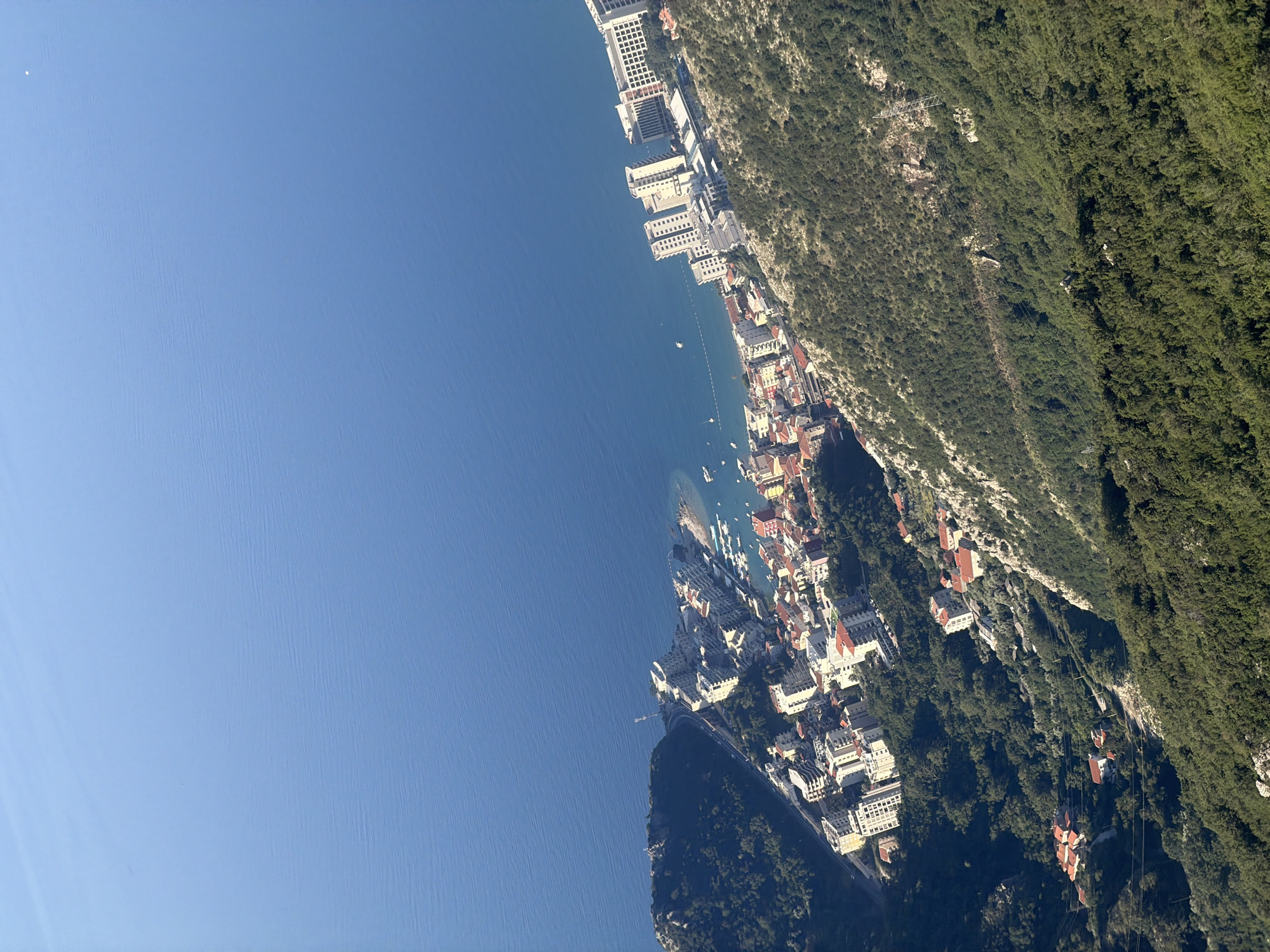 This is an aerial, high-angle, medium shot of a coastal city in Stanišići, Montenegro, situated between a lush green mountainside and the Adriatic Sea. The city features a dense collection of buildings, predominantly white and beige, with red-tiled roofs. The buildings are multi-story, with some appearing to be hotels or apartment complexes, especially those closer to the coastline. Several smaller structures with red roofs are scattered further up the mountainside, suggesting residential areas or villas.
The coastline is lined with a beach, dotted with boats and smaller vessels anchored in the calm, blue water. A paved road winds along the coast, connecting different parts of the urban area. The sea is a vast expanse of deep blue, with a slight texture indicating ripples or small waves. The mountainside is densely covered in green trees and vegetation, with rocky outcrops visible in some areas. The scene appears tranquil and bathed in natural sunlight, suggesting it was captured on a clear day. There are no people or animals visible in the image.
